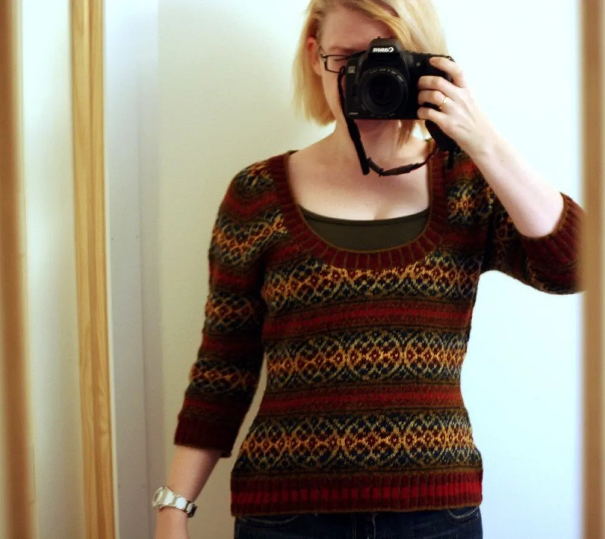 An old selfie using a black Canon camera held up in front of my face. I have light skin and short, straight blonde hair in a bob. Rectangular dark-framed glasses are visible above the camera. I’m taking a photo of the fitted knit jumper with a scoop neckline and three-quarter-length sleeves that I’m wearing. It has horizontal patterned bands in dark red, navy/dark blue, tan, and gold, with a ribbed dark red hem and neckline. A dark olive tank top is visible underneath at the neckline. Dark blue jeans are visible at the bottom of the image. A white watch is on my left wrist.