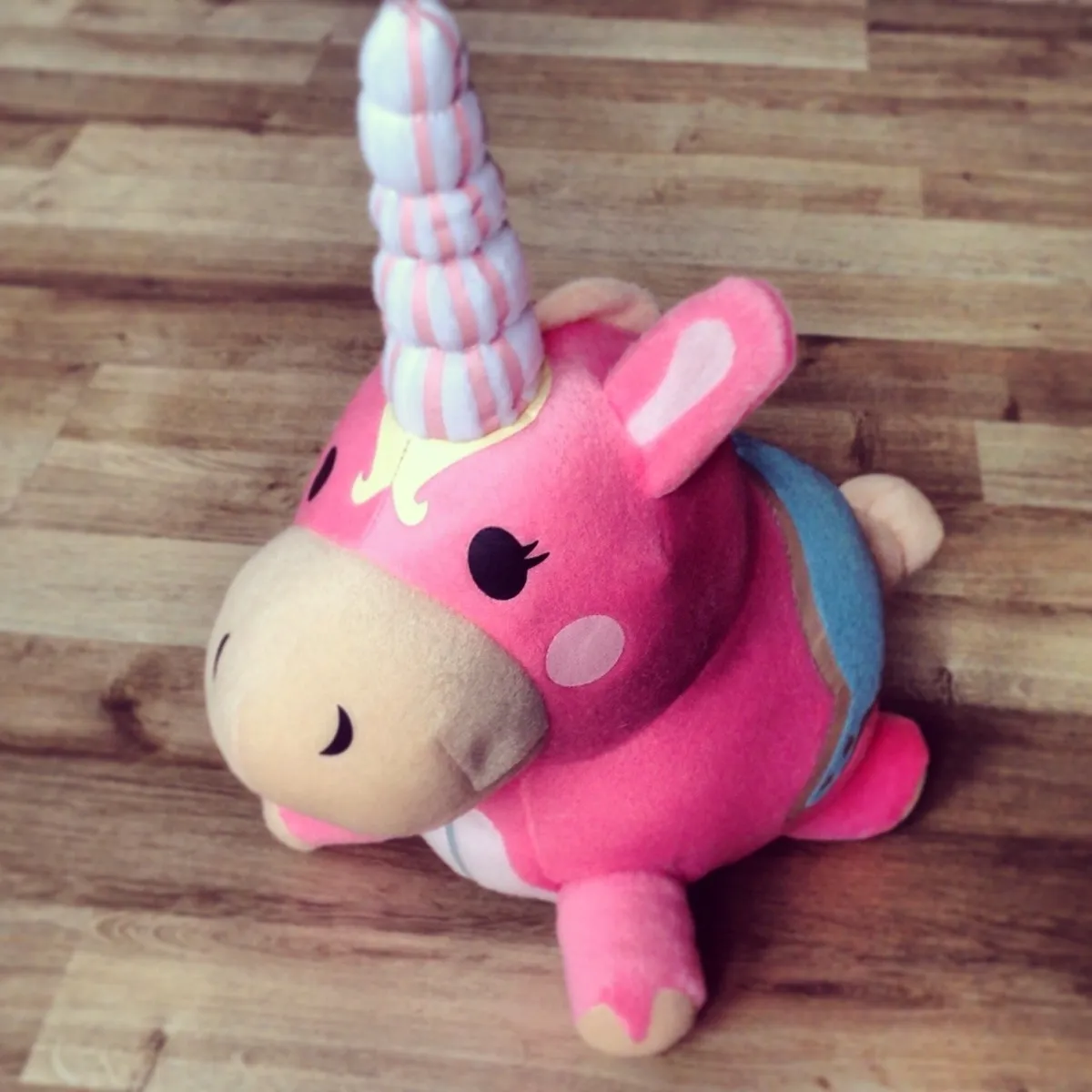 Team Fortress 2 Balloonicorn. A plush unicorn toy lying on a wooden floor. The body is bright pink with a beige snout and small black nostril marks. One visible eye is black with a small eyelash shape, and there is a pale pink circular cheek spot. A tall, spiralled horn rises from the forehead, coloured white with light pink stripes, with a small yellow decorative shape at the base. One ear is visible, pink with a lighter inner patch. The legs are short and pink with beige hooves. There is a blue section on the back, and a small beige tail is visible at the rear.