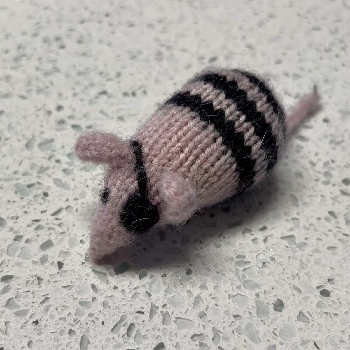 A small knitted toy mouse on a light grey speckled counter top. The mouse is mostly pale pink yarn with a pointed snout, two small rounded ears, and one black oval eyes. The other eye is obscured by the tiny black eyepatch the mouse is wearing. The back half has three black and pink alternating stripes, and a thin pink tail extends out behind it.