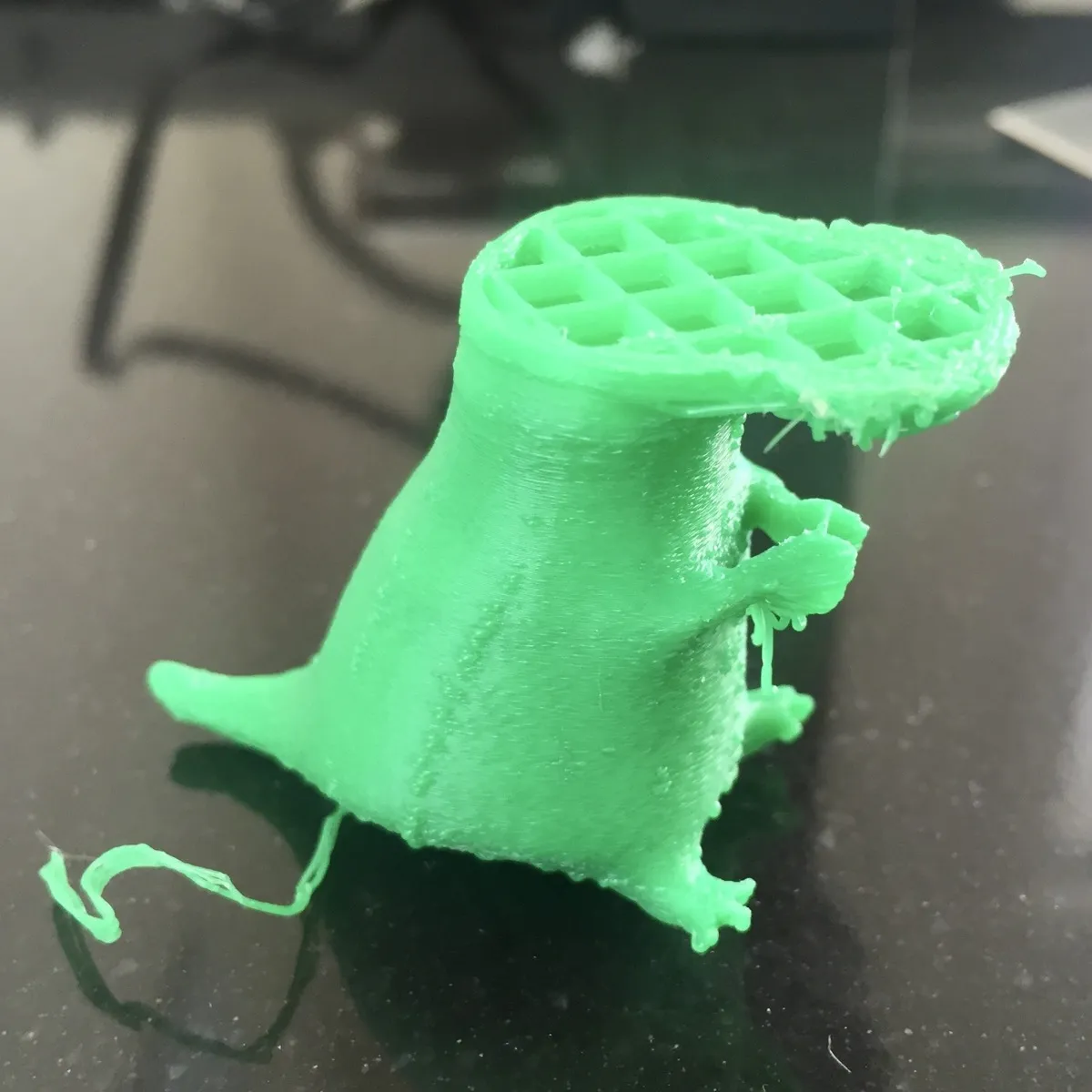 A small bright-green failed 3D-printed plastic figure on a dark tabletop. The object has a rounded, cone-like body with visible horizontal print lines, a short tail-like extension on the left, and two small arm-like protrusions on the right. The top is a circular flat area with a grid/honeycomb infill pattern exposed, and the right edge of that top surface is messy with stringy plastic and rough, torn-looking edges. There are also loose strands of filament trailing from the lower left side and some stringing around the right-side protrusions.
