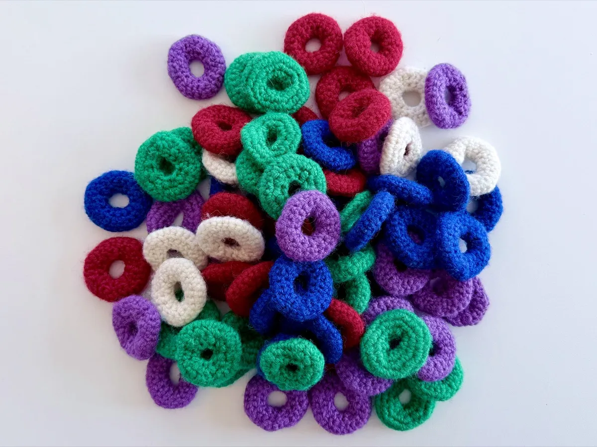 A heap of small, felted, crocheted rings on a plain light surface. The rings are all about the same size, shaped like flat donuts with a hole in the centre. They are made in solid bright colours: green, red, purple, blue, and off‑white. The rings are loosely stacked and overlapping in a heap, with no particular pattern or arrangement. The yarn looks soft and slightly fuzzy, and the stitches are tight and even, giving each ring a firm, rounded edge.