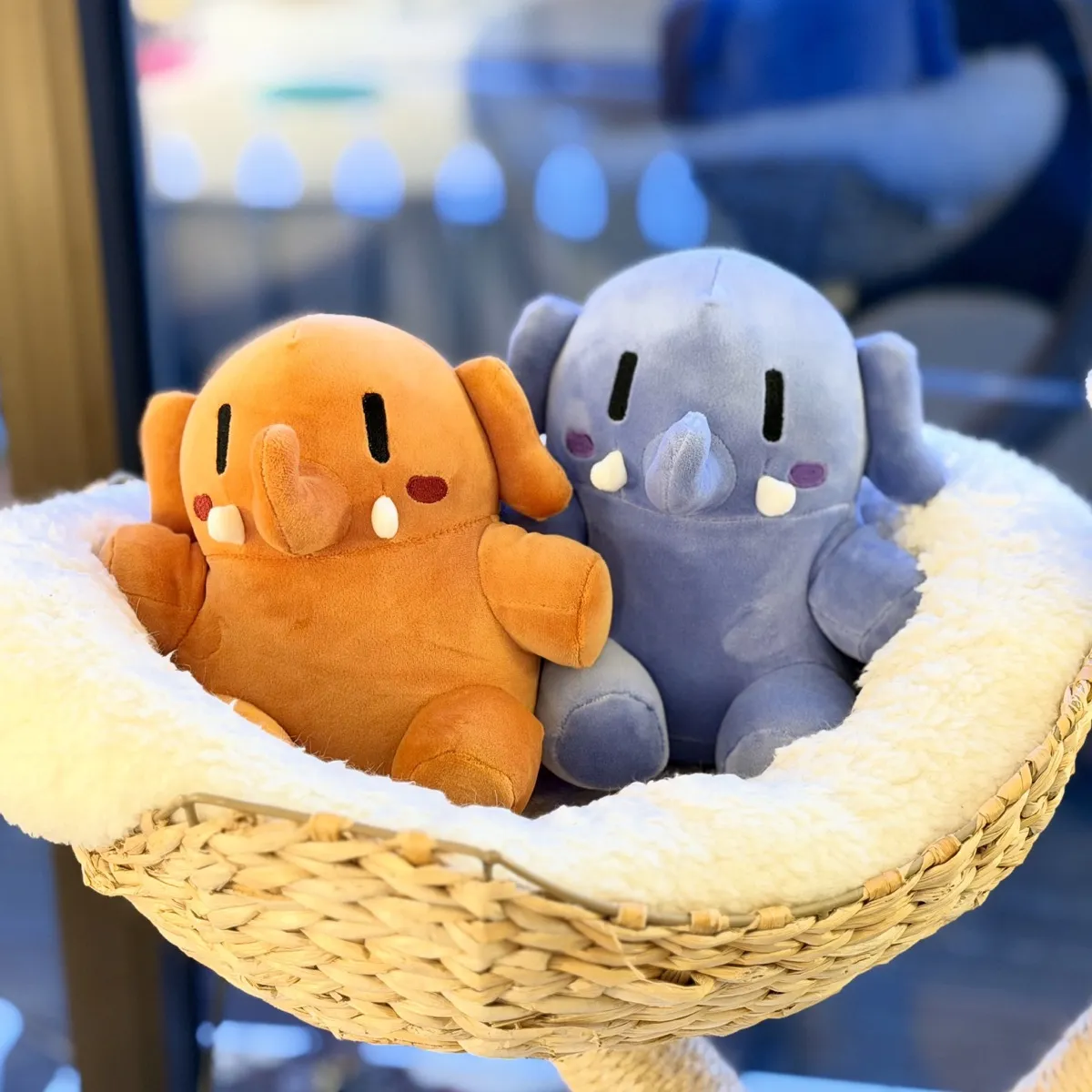 Two plushtodons (plushie elephant-like creatures), one orange and one blue, are sitting together in a soft, white, fluffy cat-tree basket. Both have rounded bodies, small arms and legs, black vertical eyes, blushed cheeks, and little white tusks. They are V smol and incredibly cute!