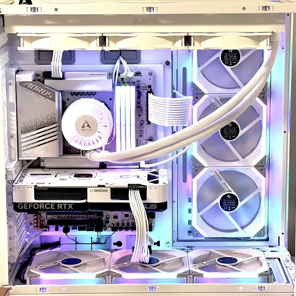 A white desktop PC interior with white components, white sleeved cables, and RGB lighting in pastel blue and purple. A white ARCTIC liquid CPU cooler is mounted on the motherboard, with a round ARCTIC pump head near the centre-left and two white braided coolant tubes running to a radiator at the top. Three white fans are mounted on the top radiator. On the right side, three vertically stacked white case fans are mounted at the front/side intake area. In the middle, four white RAM sticks with lighted strips stand upright. A large white graphics card is installed horizontally; the side reads “GEFORCE RTX” in large letters, and “ASUS” is also visible. At the bottom, three more white fans are mounted on the case floor. The motherboard’s left-side heat sink cover shows “AORUS” and “TEAM UP FIGHT ON.” The word “ARCTIC” is also visible on the top radiator area and on the pump head.