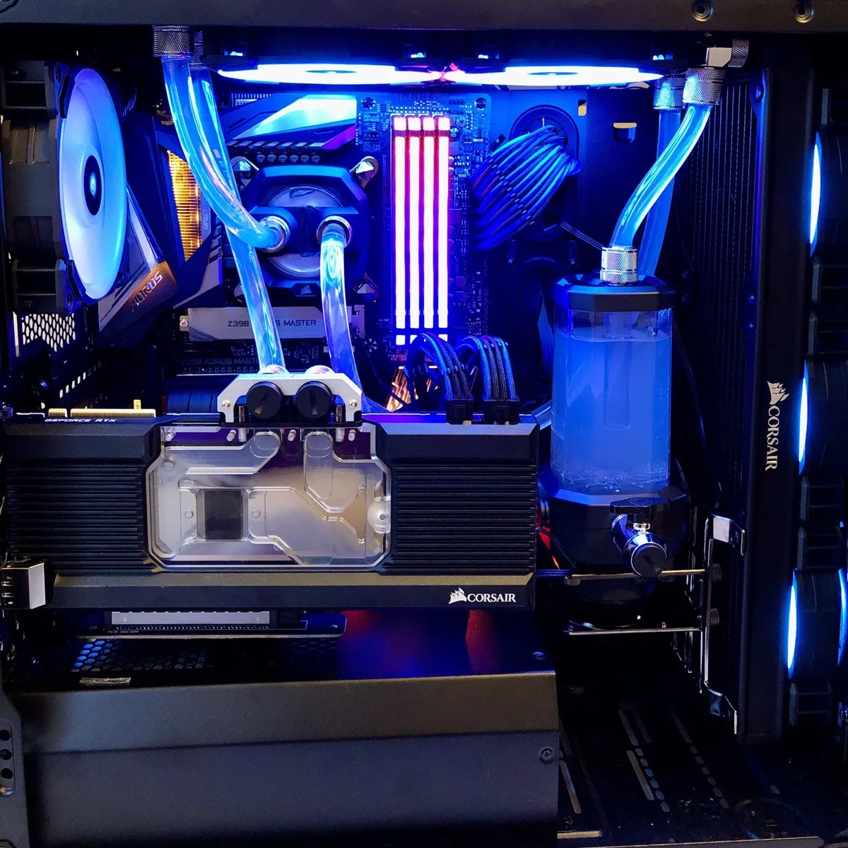 A custom desktop computer interior with liquid-cooling components and blue lighting. Clear tubes run from a CPU water block near the centre-left to a cylindrical reservoir/pump on the right and to a radiator area on the right side. Two bright, horizontal lighted fans are mounted at the top. A large graphics card with a clear water block cover spans across the lower middle; “CORSAIR” is printed on it in white, and “GEFORCE RTX” is visible on the left edge of the card. Two vertical RGB RAM sticks glow in the centre (pink/purple and white). On the left, a large white case fan is visible, and “AORUS” can be read on a component near it. On the right side, a vertical radiator or fan stack shows “CORSAIR” printed in white. The reservoir contains blue-tinted coolant with small bubbles visible near the bottom.