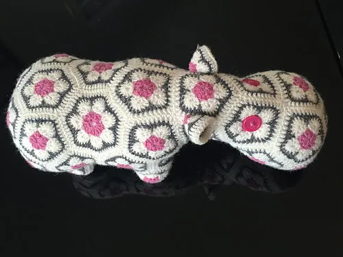 A crocheted stuffed hippopotamus toy with a white yarn body covered in repeating flower-like hexagon motifs outlined in dark gray, with pink centers and white petals. The hippo has small rounded ears, a short tail, four stubby legs, and two bright pink button eyes. The snout is large and rounded, with the same floral pattern continuing across it.