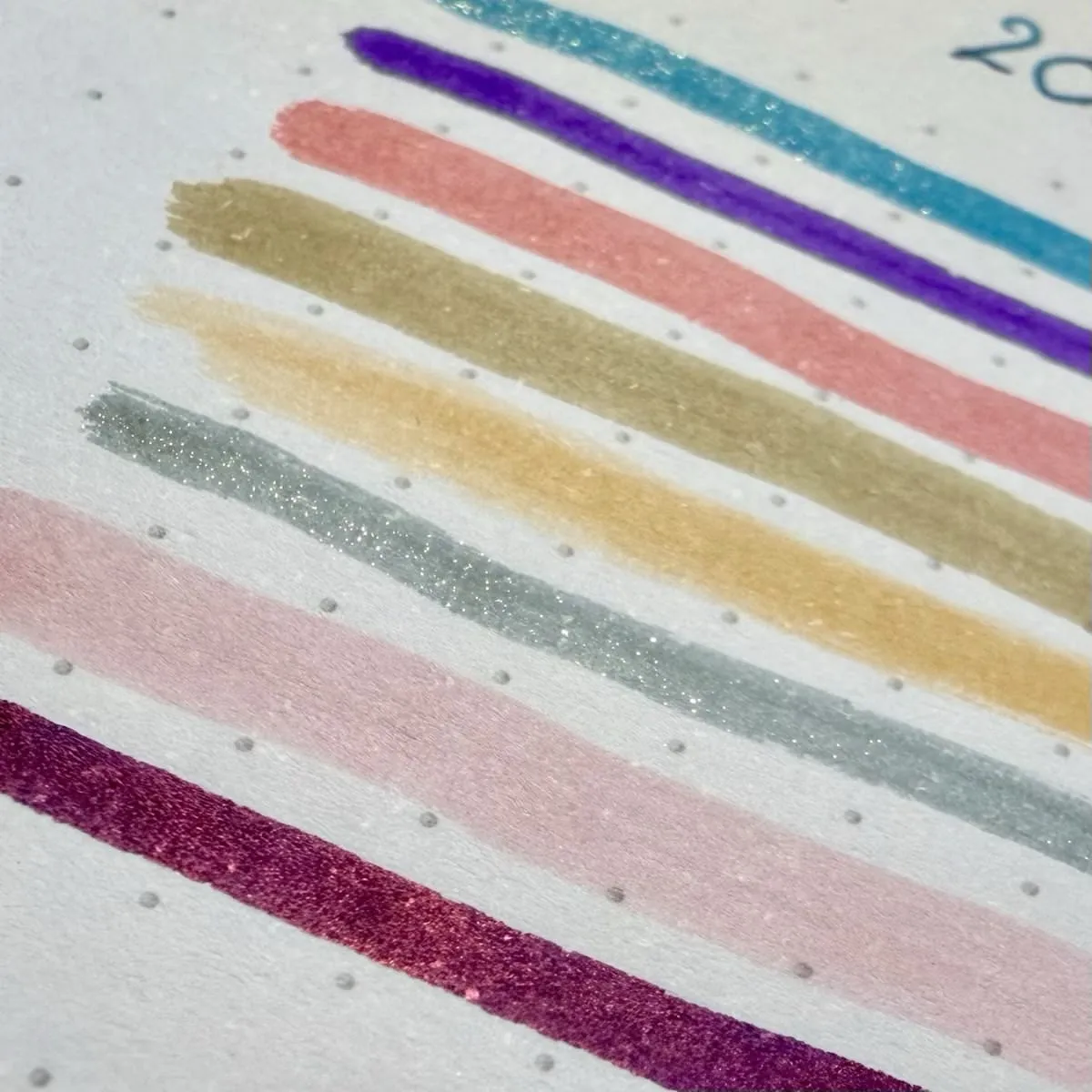 Colourful ink swatches on Rhodia dotted paper, shown in horizontal strokes. The swatches include bright and pastel colours such as teal, purple, salmon pink, antique gold, orangey yellow, grey-green, pale pink, and reddish-purple. Three inks appear shimmery or metallic under the light, and individual glitter particles are visible. The image is a close-up, highlighting the glittery and colour-shifting nature of the ink.