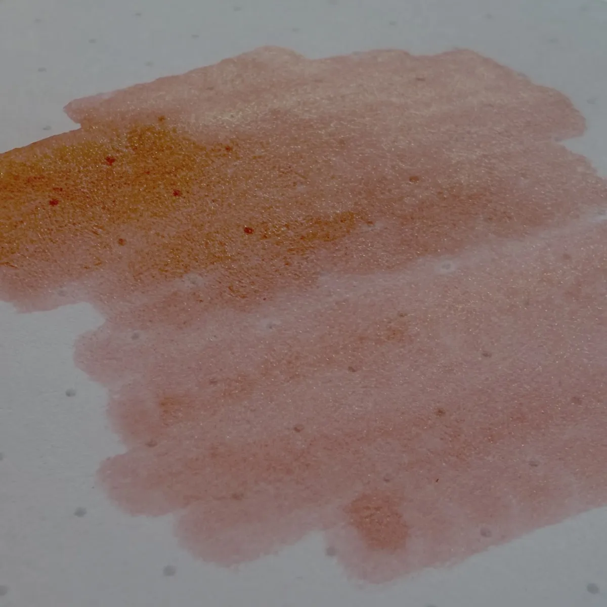 A swatch of orangey red ink on dotted white paper, darkest at the top and fading towards the bottom. The darkest area has visible sparkle and shimmers with an orange glow.