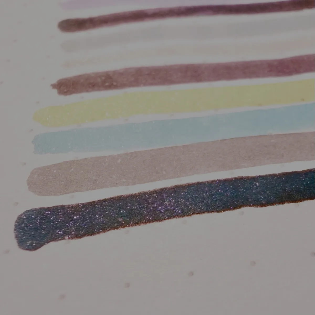 Coloured ink swatches on dotted Rhodia paper. There are several horizontal lines in different colours, including purple, gray, brown, yellow, light blue, taupe, and a dark shade with visible multicoloured glitter. The lines are drawn thickly, and the shimmery one at the bottom catches the light, showing sparkles in its ink.