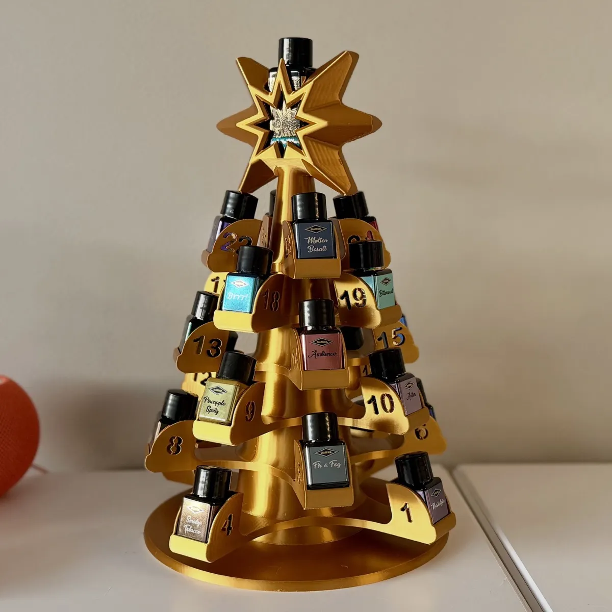 Golden 3D-printed tree-shaped advent calendar holder for the Diamine Inkvent bottles. The tree has numbered shelves from 1 to 24, each containing a small bottle of ink. The tree is topped with a star decoration, which also holds the large bottle revealed on the 25th.