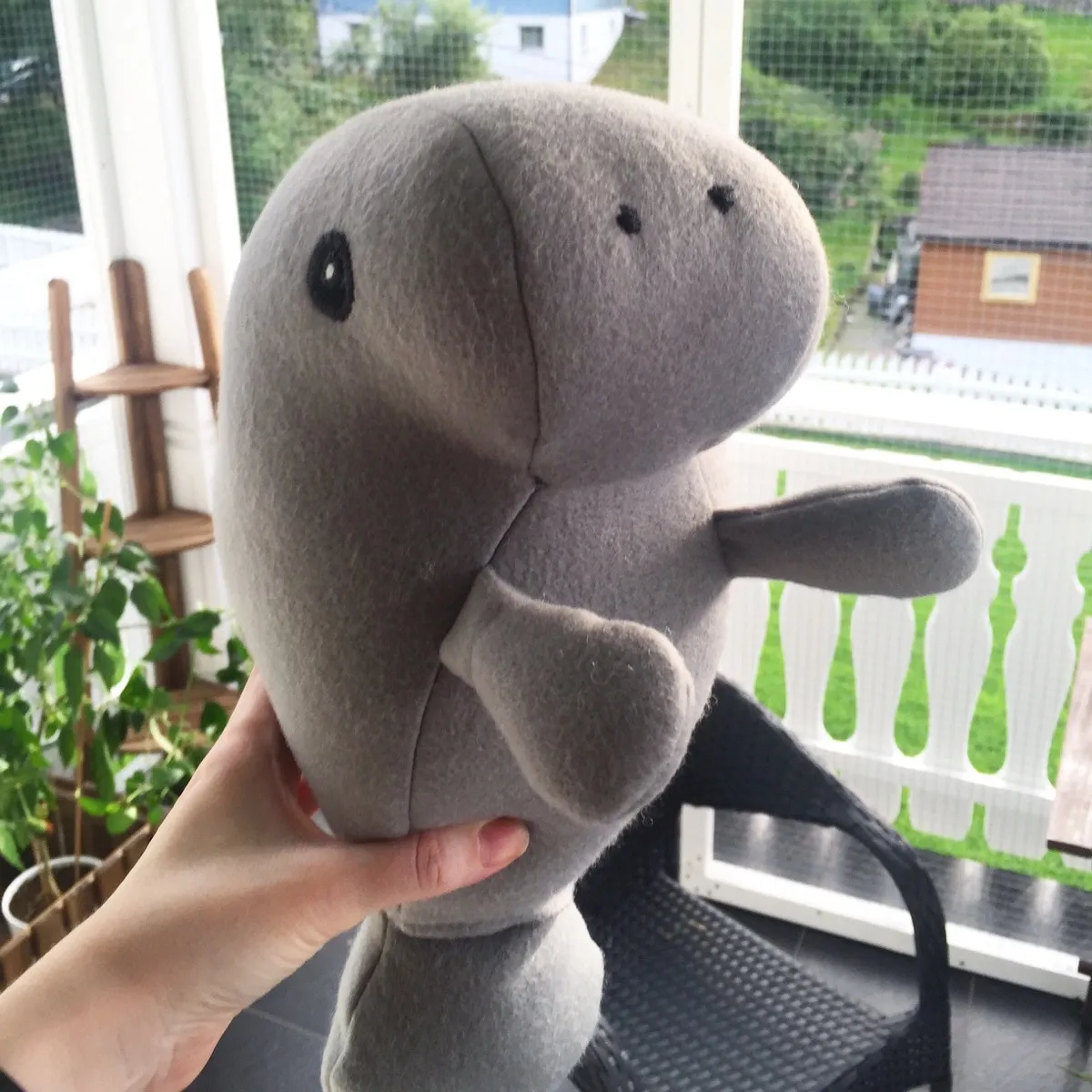 A grey manatee plush toy shown in profile with a large rounded snout. The toy has one shiny black oval eye and two small black dots on the snout that resemble nostrils. It has two small flipper-like arms, one pointing forward and one downward, and a rounded body with visible seam lines running down the face and along the side. The fabric looks soft and slightly fuzzy. In the background there are bright windows, green plants, a wooden stand, and an outdoor view with grass and a small building.