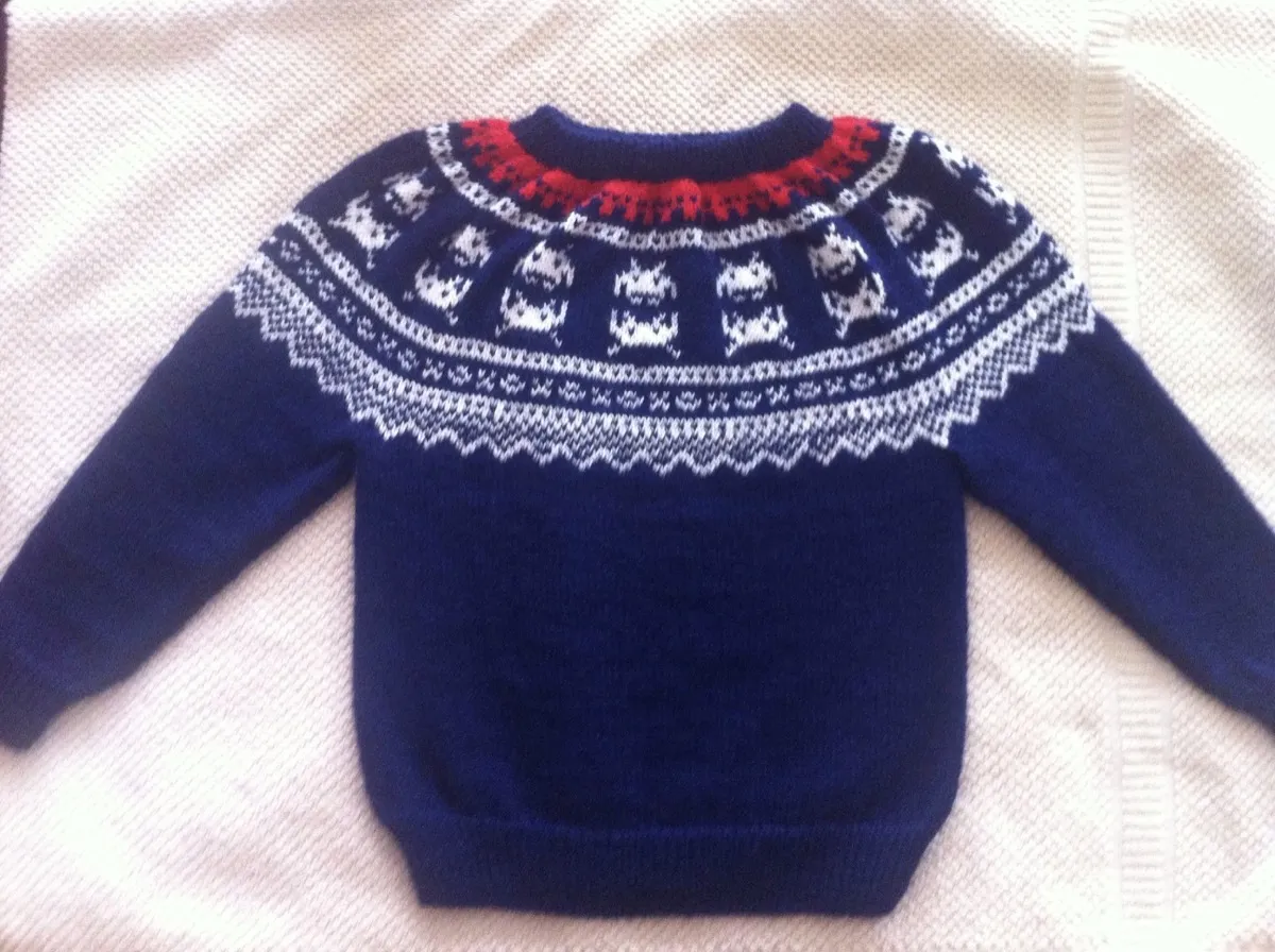 A navy-blue knit baby jumper laid flat on a light-coloured towel. It has long sleeves and a round crew neck. It is knit in a traditional Norwegian Marius pattern and colours (navy body, with white Fair Isle pattern across the yoke, and a smaller band of red pattern near the neck), but part of the pattern on the yoke has been swapped out for Space Invader designs instead. The cuffs and bottom hem are ribbed.