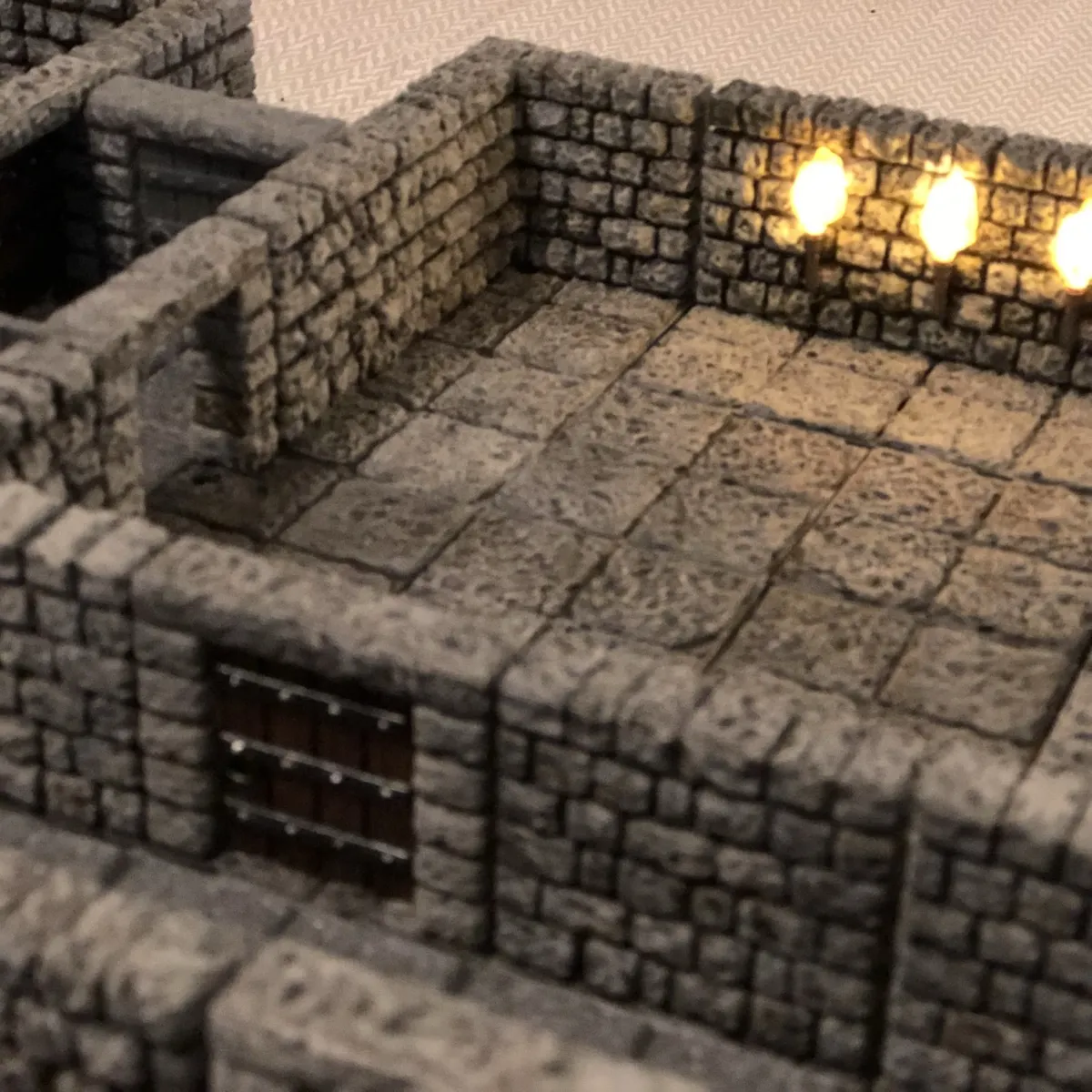 3D printed mini terrain for D&D. A small stone-walled miniature room with a tiled stone floor and three lit wall torches. The walls are made of gray, blocky “stone” bricks, forming a rectangular room. The floor is made of square stone tiles in a grid, with darker grout lines and speckled texture. On the back wall, three torches are mounted close together, each with a flame-shaped light, that is brightly lit. Along the lower wall there is a small dark doorway or gate with horizontal metal-like bars. At the upper left, a separate stone structure creates a narrow passage or alcove leading into a darker recessed area.