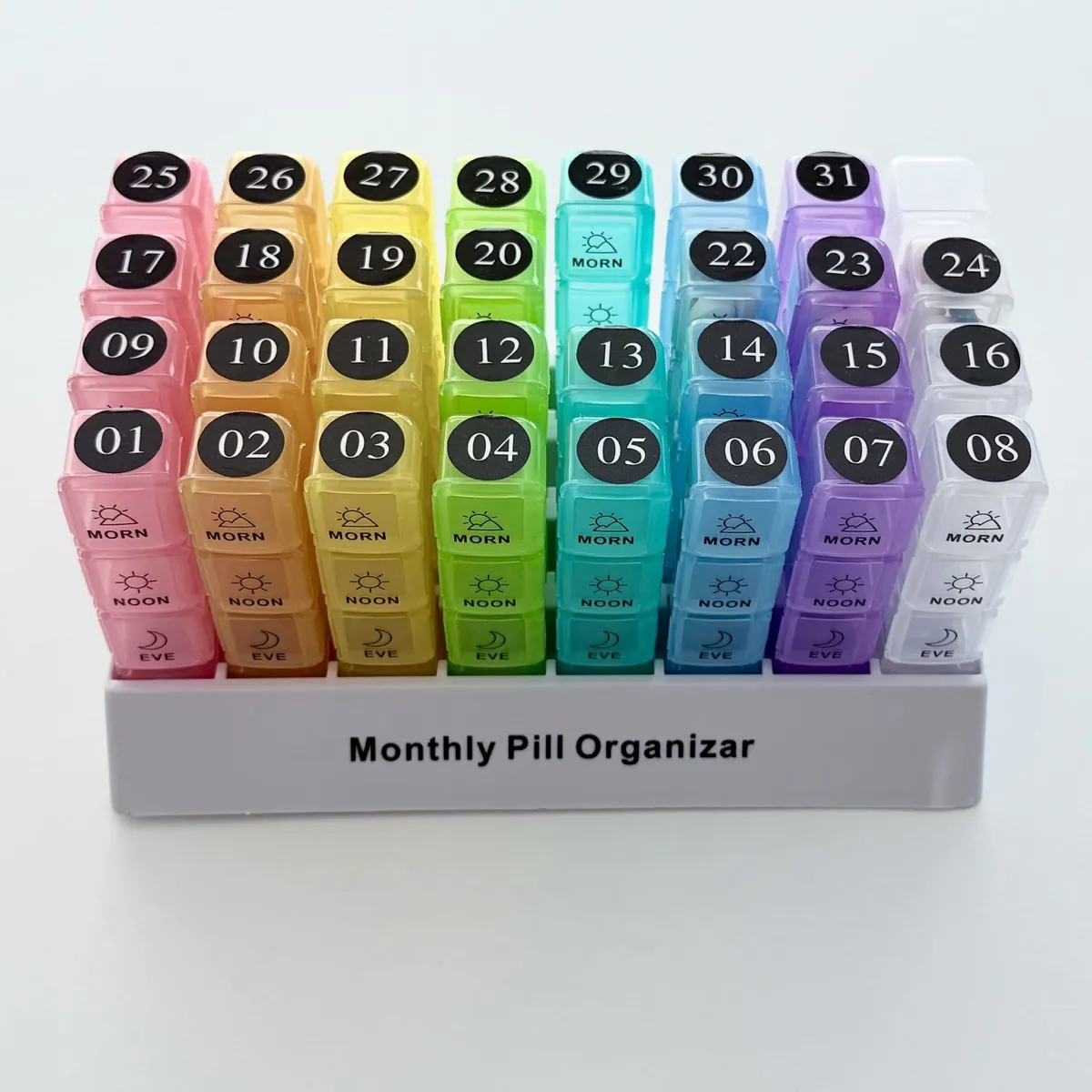 A monthly pill organiser with 31 detachable daily pill boxes arranged in a row on a white base. The boxes are translucent and color-coded in a gradient from pink (left) to orange, yellow, green, teal, blue, purple, and clear/white (right). Each daily box has four stacked compartments labeled with time-of-day words and icons: MORN (sunrise icon), NOON (sun icon), EVE (crescent moon icon), and a fourth compartment hidden by the base. A round black sticker with the day of the month number in white is affixed to the top of each individual pill box (numbers 01-31). Pill box number 21 is missing, and the clear pill box in the top right corner does not have a sticker. Text in black on the base reads: Monthly Pill Organizer.