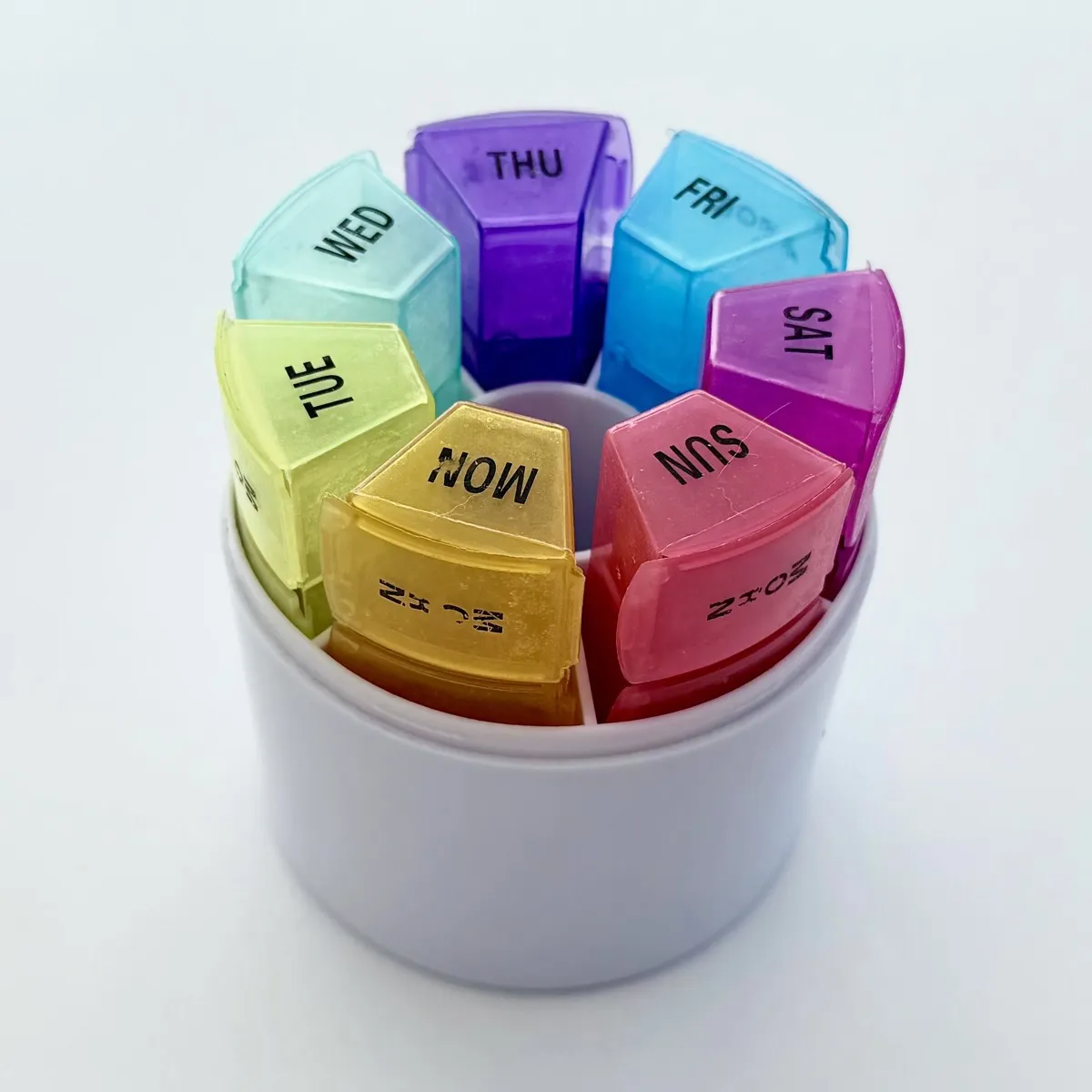 A round pill organizer with seven translucent, wedge-shaped compartments arranged in a circle inside a white cylindrical base. The compartments are different colors and each lid is labeled with a day of the week. Visible text on the lids from the top and going clockwise: THU, FRI, SAT, SUN, MON, TUE, WED.