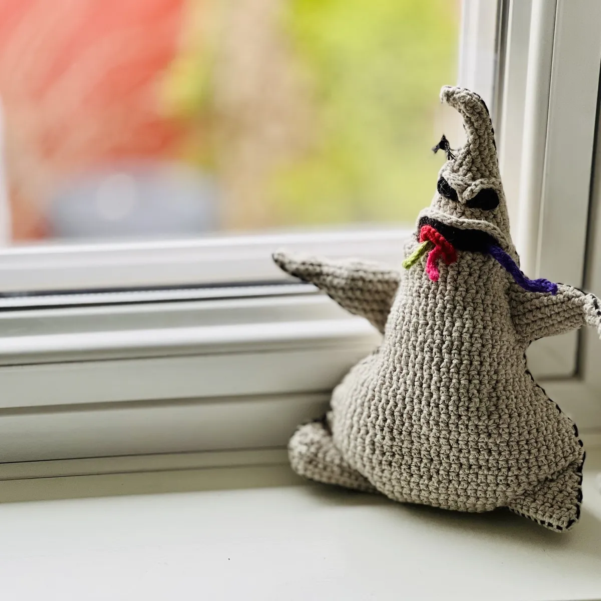 A small(ish) crocheted doll of Oogie from The Nightmare Before Christmas. He is posed against a window, sitting on the sill with the view outside blurred by depth of field. A small crocheted spider hangs from the top of his head and his slightly open mouth has crocheted worms of various colours spilling out.