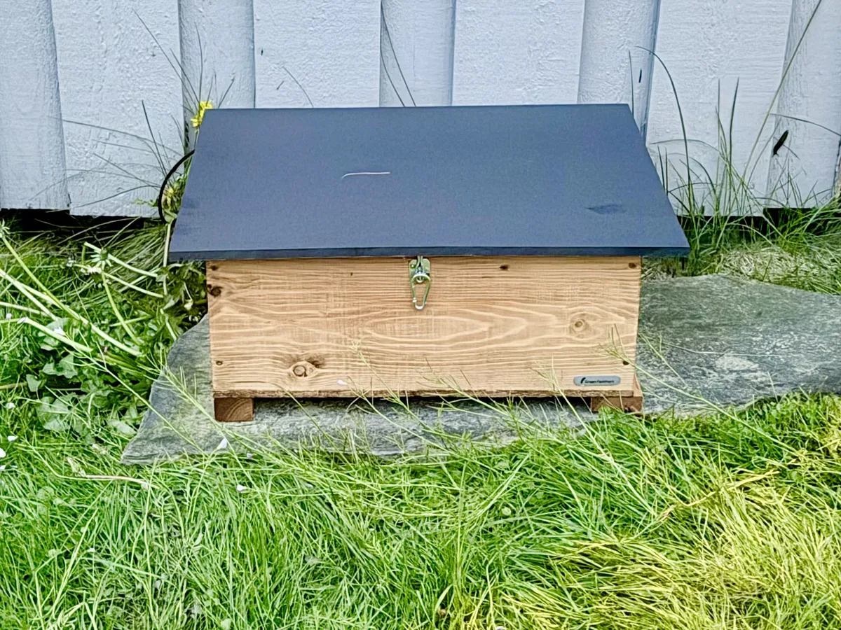 Piggiewinkuhl Mansion A wooden hedgehog shelter with a rubberized roof resting on a stone slab, positioned against a pale grey wooden wall. The slate is surrounded by grass.