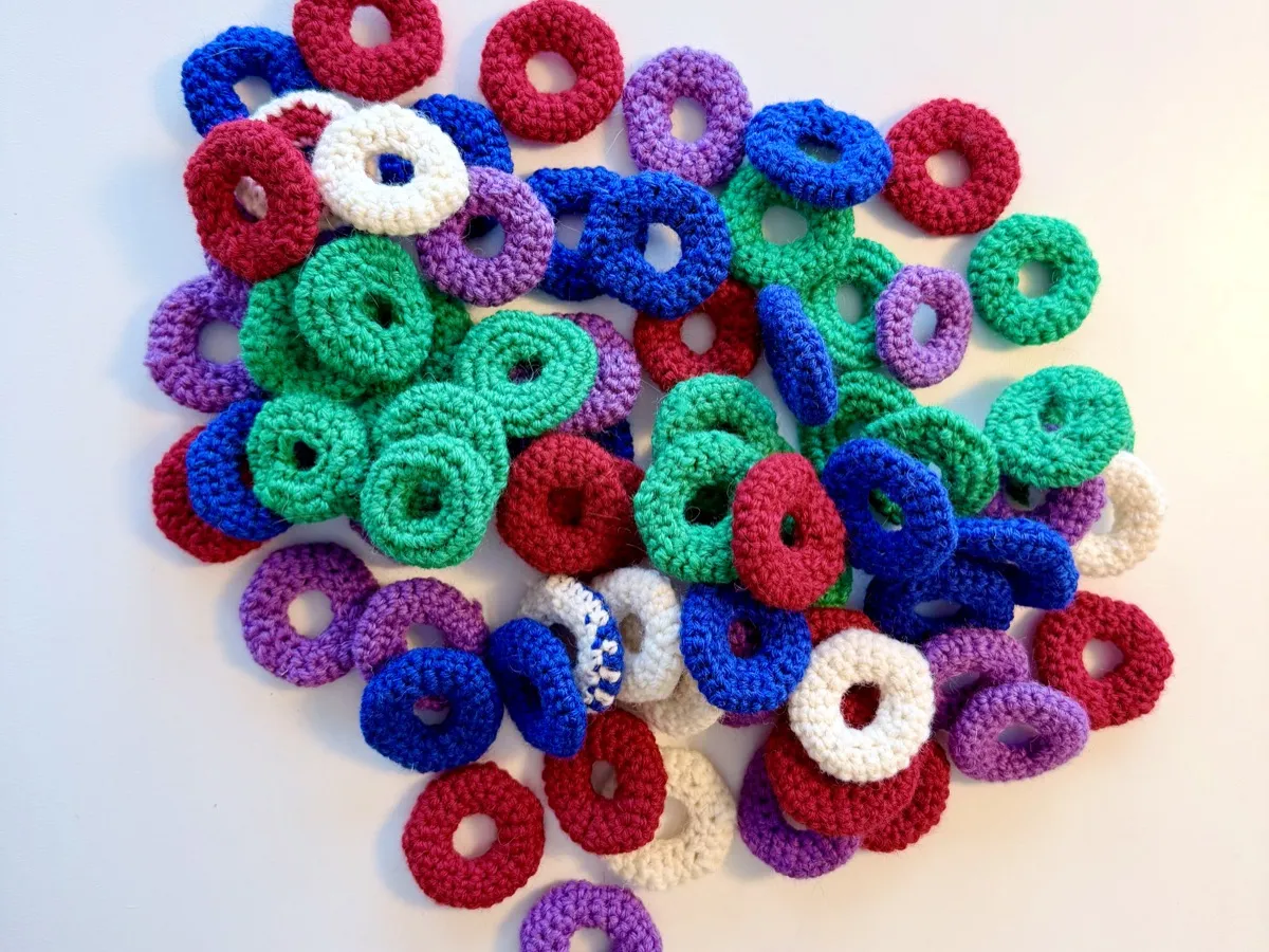 A pile of small, unfelted, crocheted rings in bright colours. The rings are flat, donut-shaped circles made from yarn. The colours include red, royal blue, bright green, purple, white, and a half blue and half white one. They are scattered randomly on a light-coloured surface, overlapping each other in a loose mound. Each ring has a slightly textured, stitched pattern and a hole in the centre about a third of the ring’s overall width.