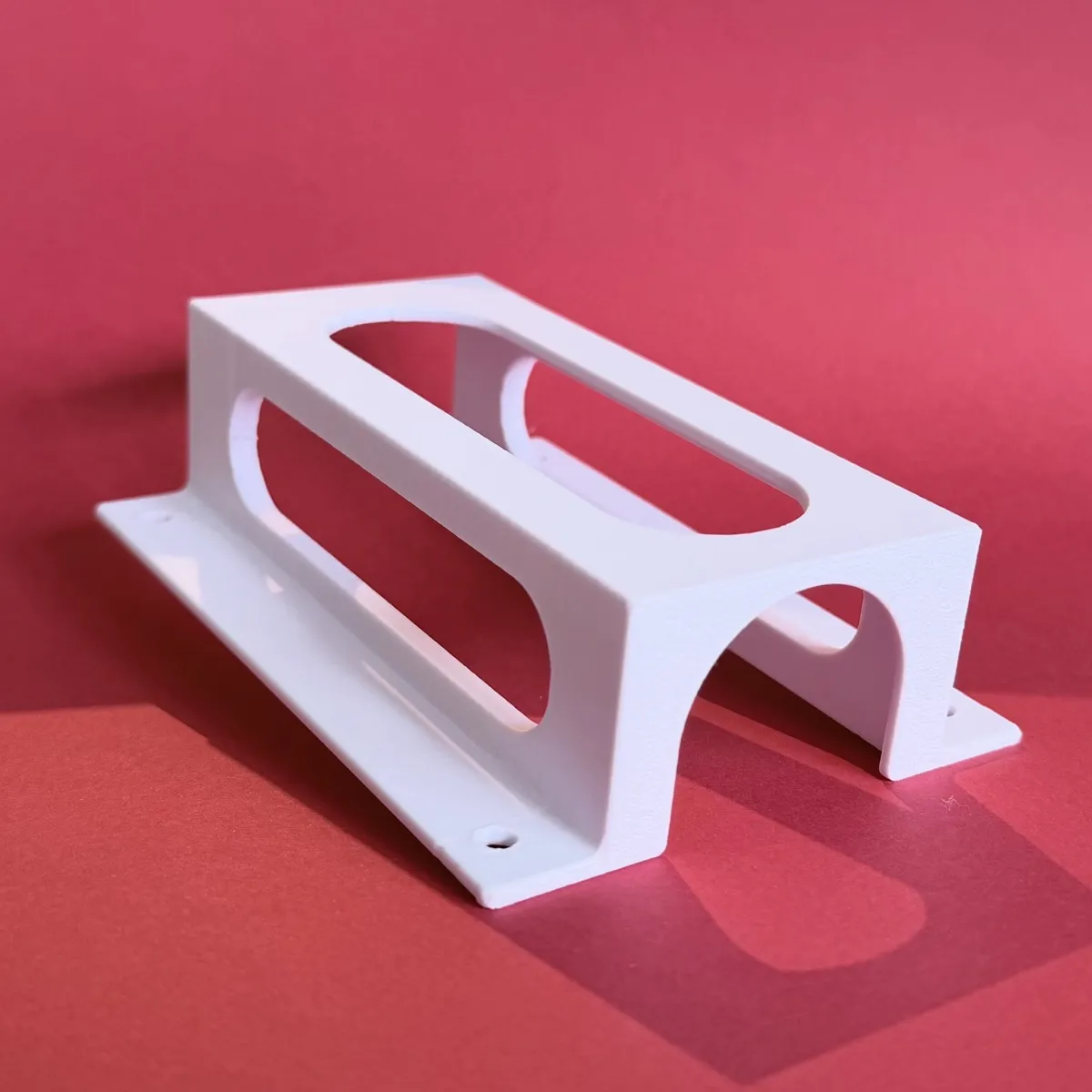 A white 3D printed rectangular bracket against a red background. On the top, a long rounded-rectangle slot runs lengthwise. Each side has a similar large rounded cutout, the top is open, while the bottom has an arched opening. Two screw holes are visible in the baseplate (one near the top, the other at the bottom).