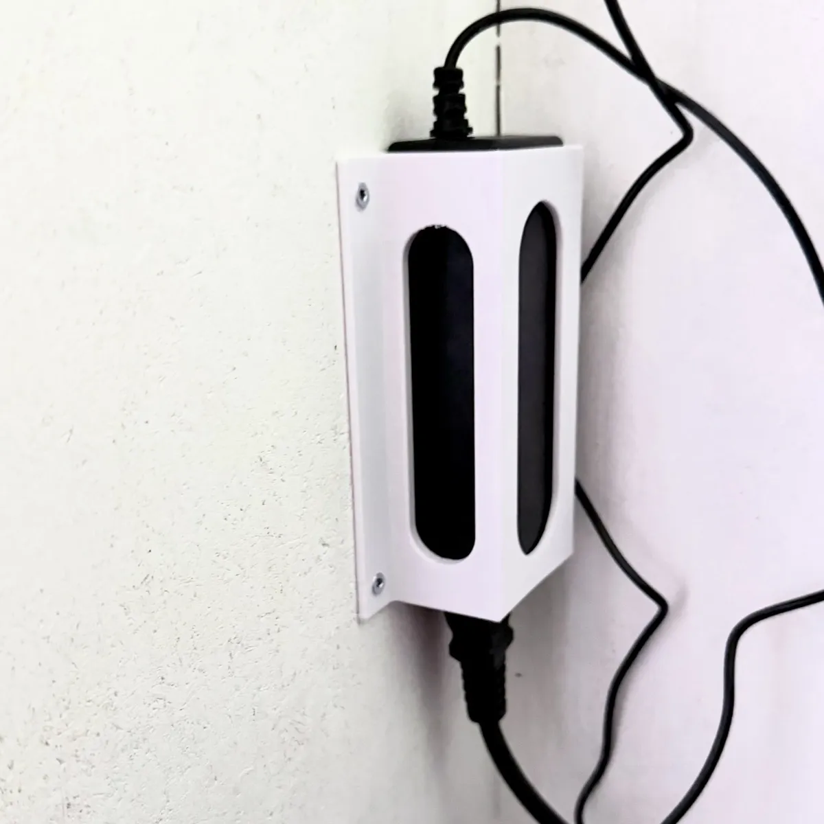 A white 3D printed wall mount holding a power brick installed near an internal corner of two white walls. Various black cables are visible, a thick one attached to a plug at the bottom of the power brick, a thinner, longer one attached to the top. 