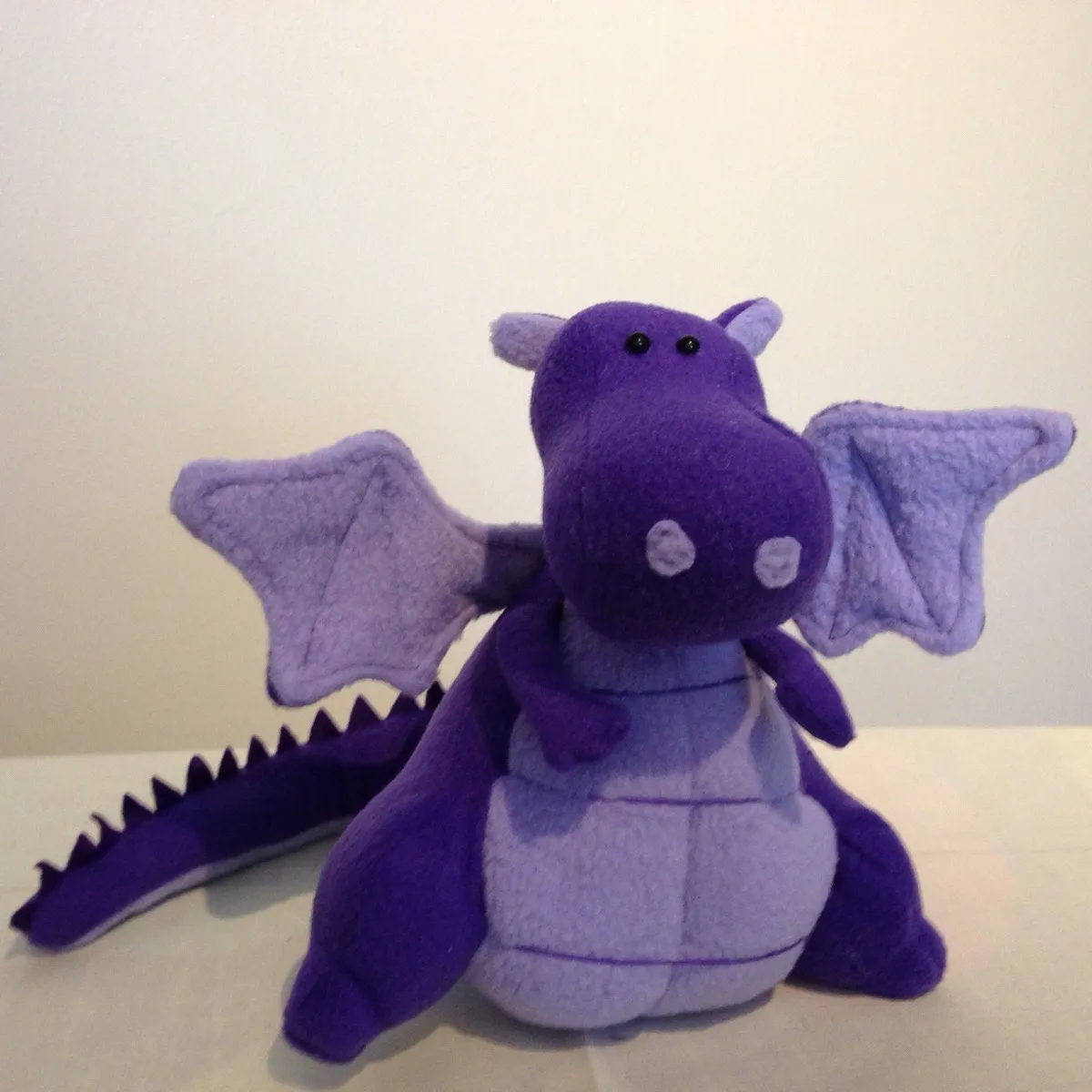 A purple plush dragon toy sitting upright on a light-coloured surface against a plain off-white background. The dragon has a rounded snout with two small light gray circles near the front, two small black bead eyes on top of the head, and small rounded ears. Two light purple wings spread out to either side with stitched vein lines. The belly is a lighter lavender colour with stitched panel lines. A long tail extends to the left with small dark purple triangular spikes along the top, and the legs are splayed forward in a seated pose.