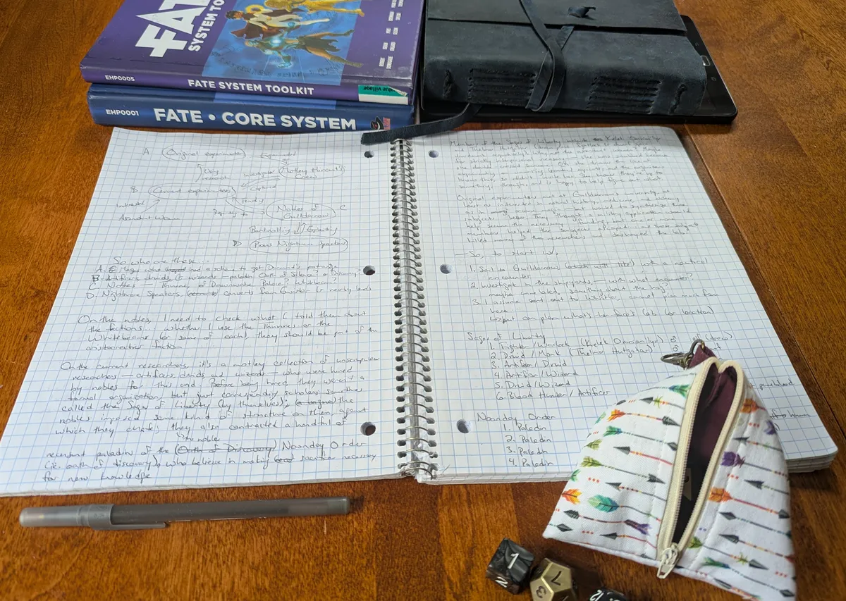 photograph of ttrpg accoutrements, including an open notebook, a closed notebook, two Fate Core books, a bag of dice, and a pen