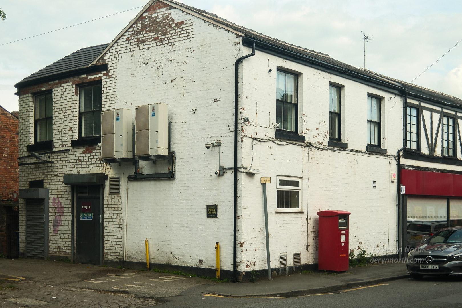 A corner-view of a building painted white. It has two large air conditioning units on the external wall and a post box outside and a car parked on the street. The building looks untidy and scruffy. 