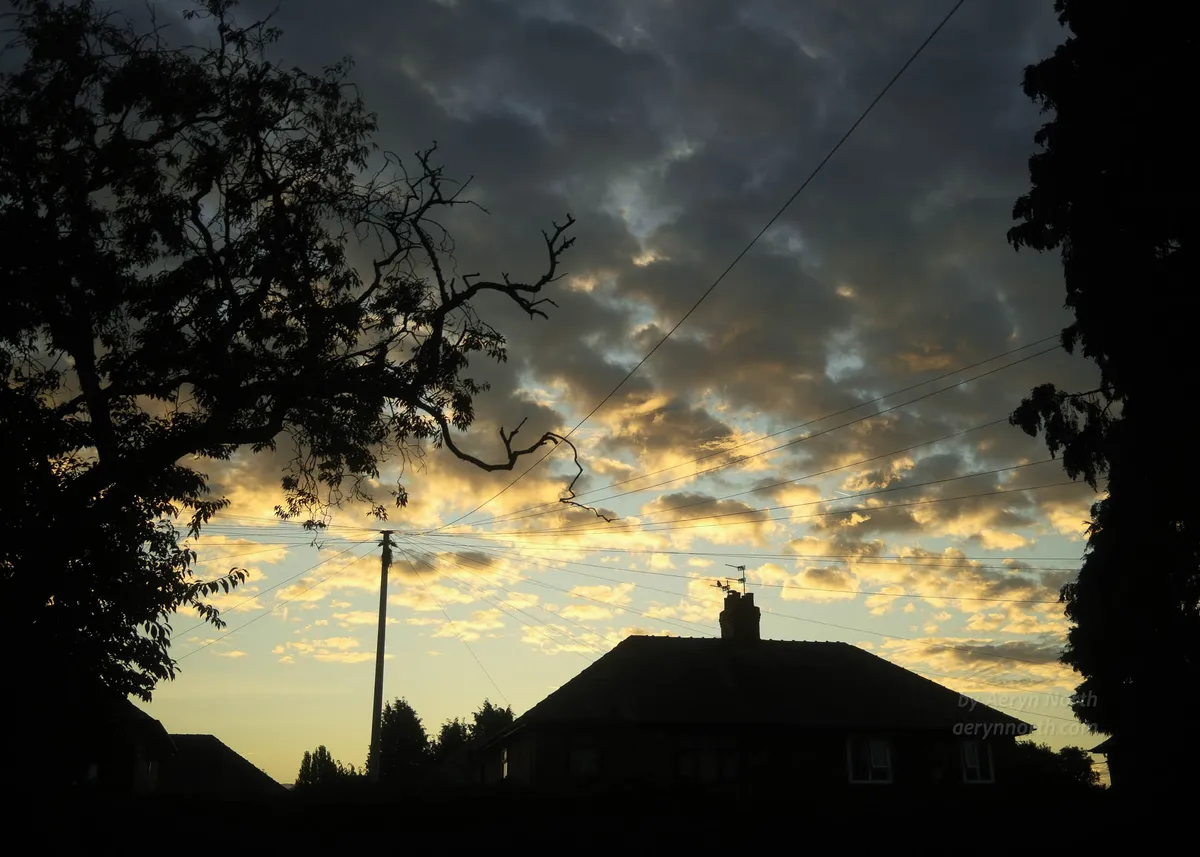 A sky filled with clouds backlit in warm colours by the rising sun is framed by a silhouetted house and trees. Telephone wires radiate from a pole.