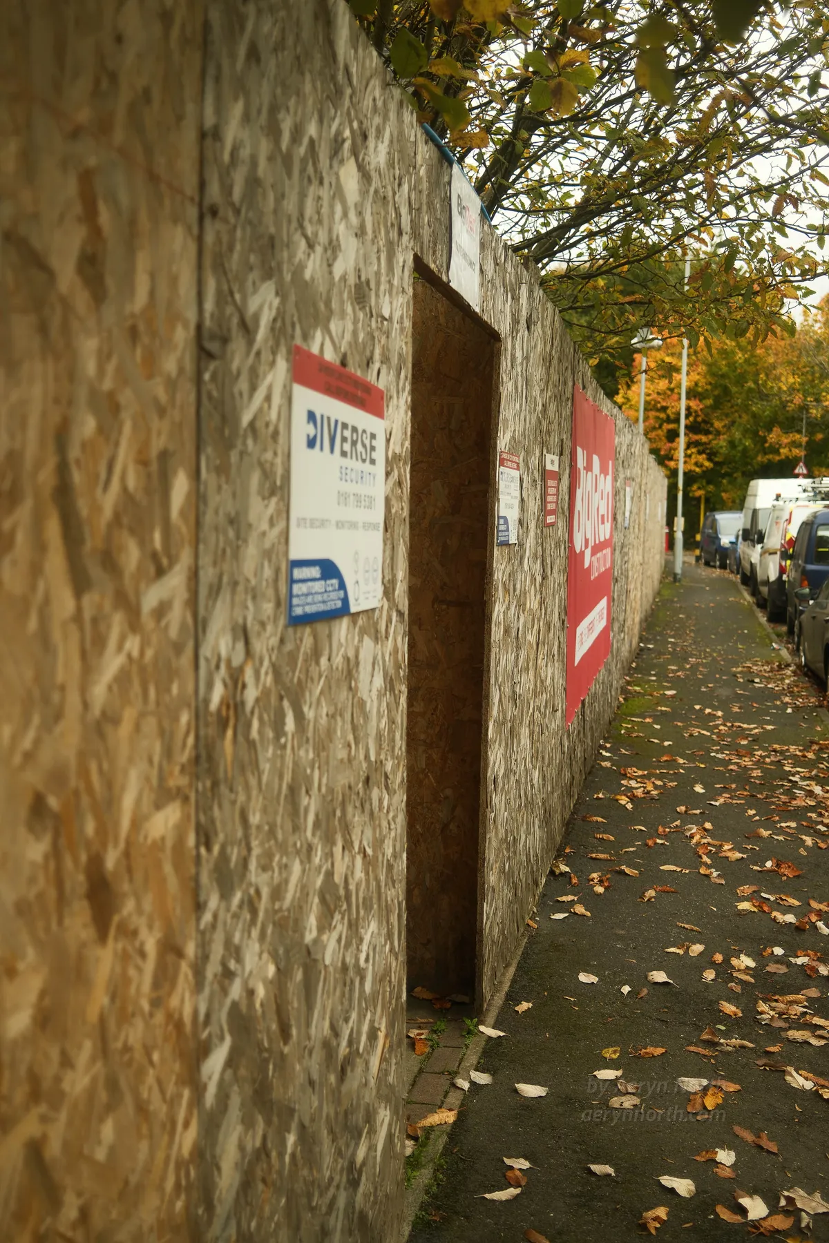 A construction hoarding runs on the left side of the street with a darkened opening. Cars are parked along the kerb and autumnal leaves are strewn on the ground. 