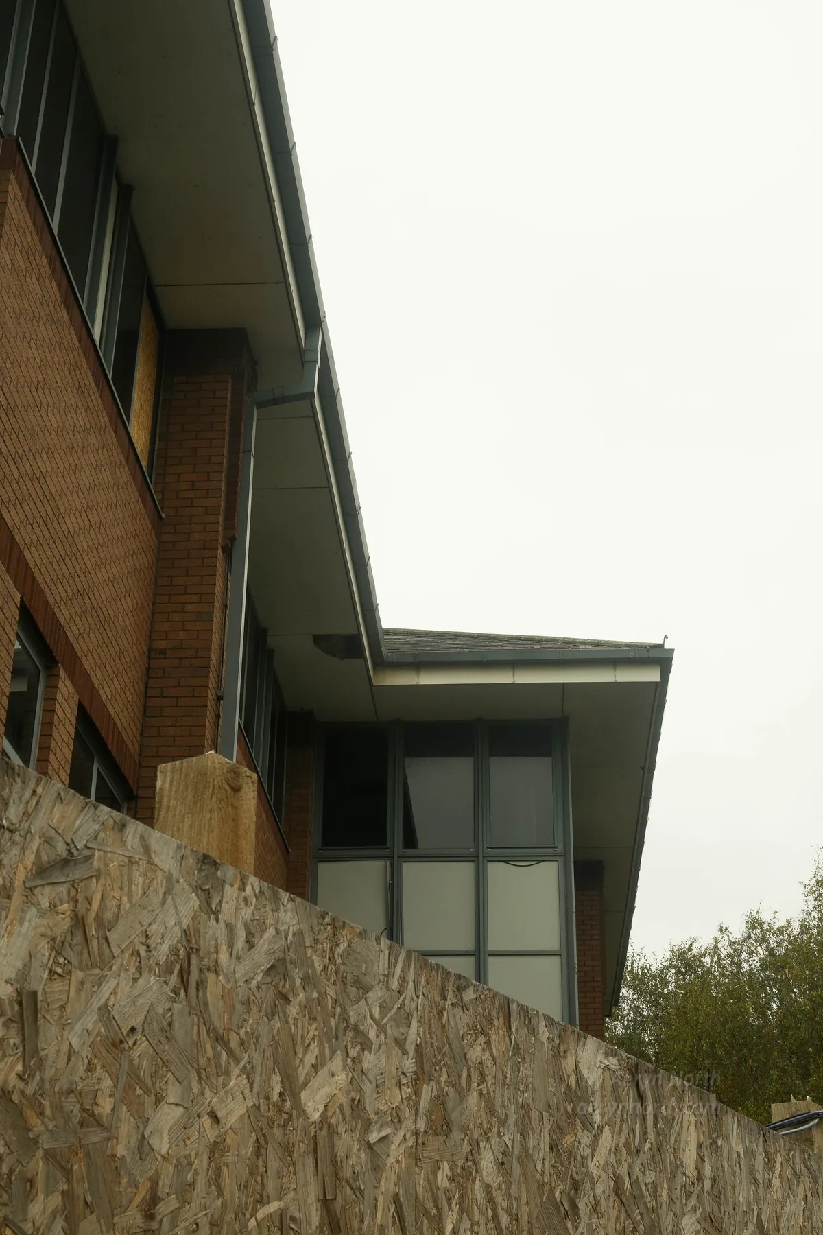 An upward view of the top of an office building, over a costruction hoarding. A featureless white sky above.
