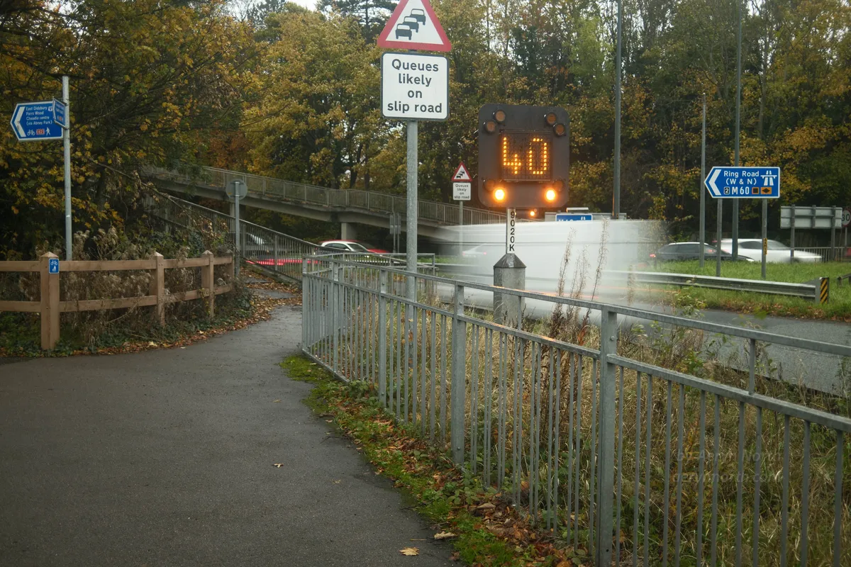 Motorway signs at a busy roundabout say Queues likely on slip road, and a large dot-matrix sign  in orange lights shows a 40 speed limit. A white van is shown exiting the roundabout. It and other cars all have motion blur. In the foreground are railings, undergrowth and a footpath. 
