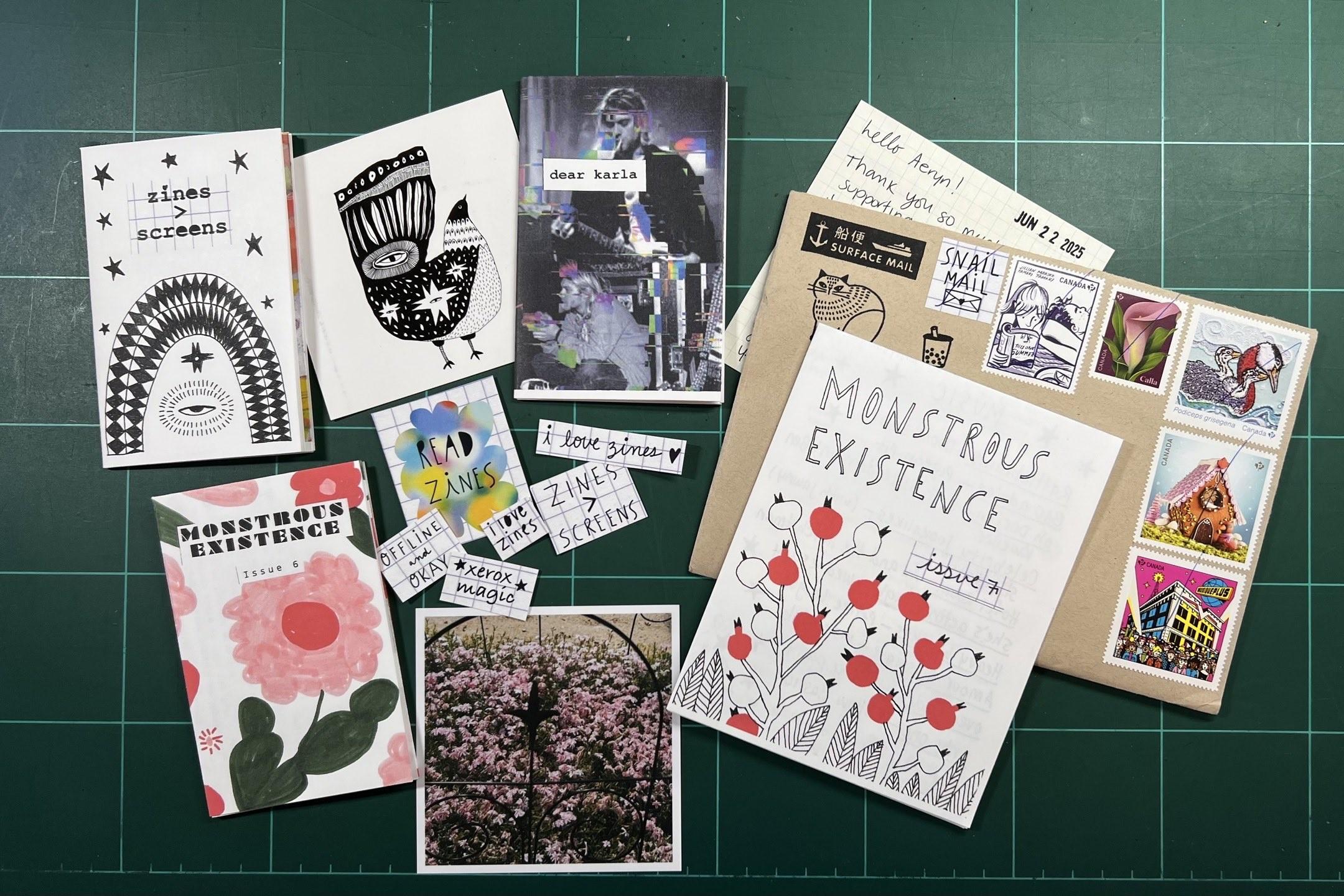 A collection of zines, a photo, an envelope with Canadian stamps, and printed materials arranged on a green cutting mat.