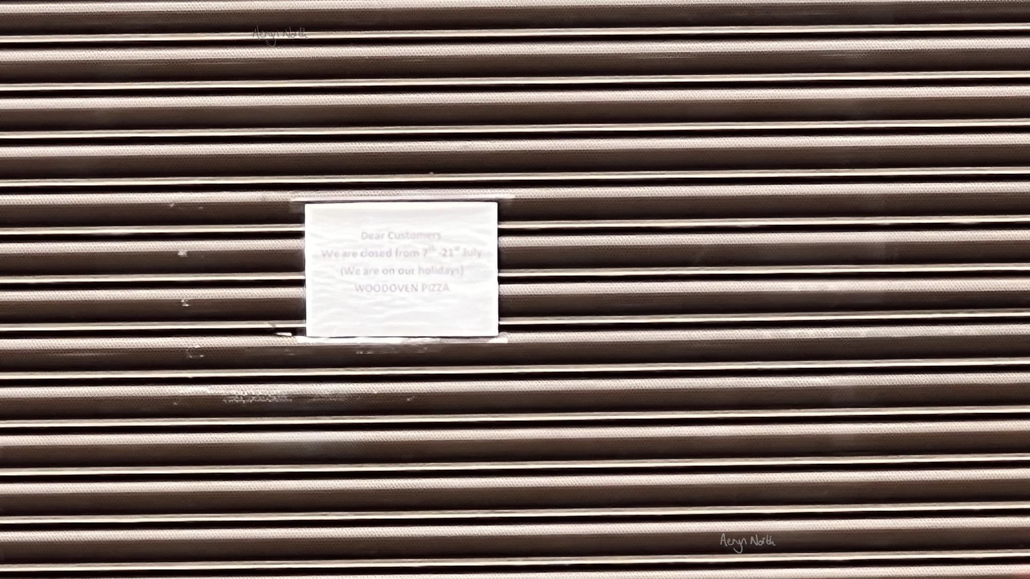 A close-up view of a closed, metal roller shutter with a rectangular white note attached to it. The note reads: Dear Customers, We are closed from 7th - 21st July (We are on our holidays) WOODOVEN PIZZA