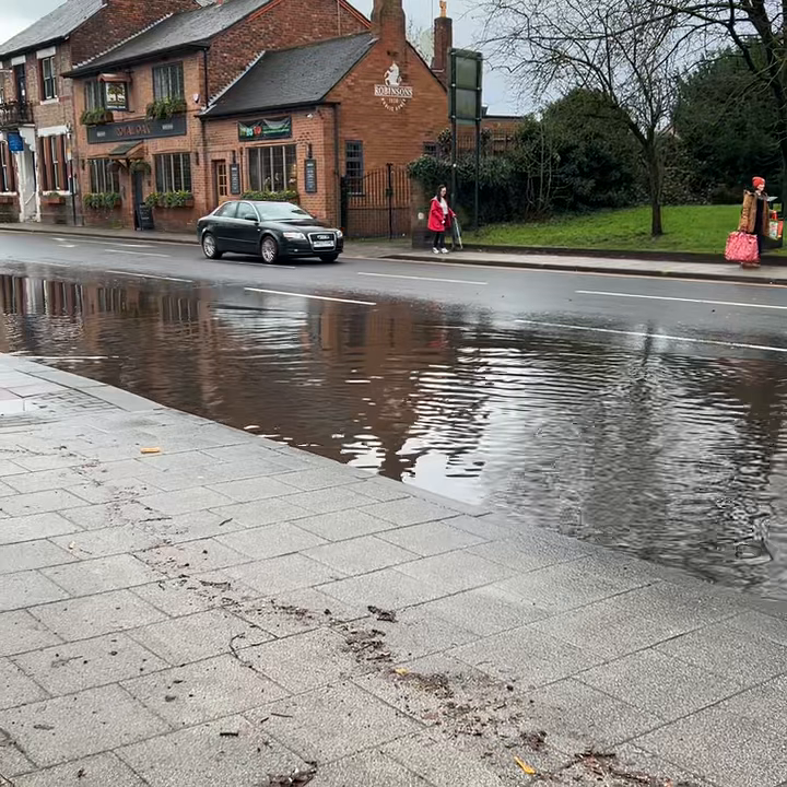 Still image from a video showing a very large puddle on a road with a red brick building. A car and pedestrians can be seen.