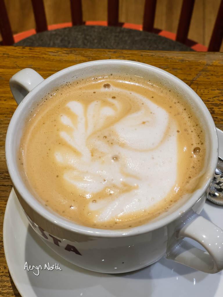 A top-down view of a Latte from Costa Coffee in a large two-handle mug. The latte foam has a pattern that looks like an animal holding some vegetation.