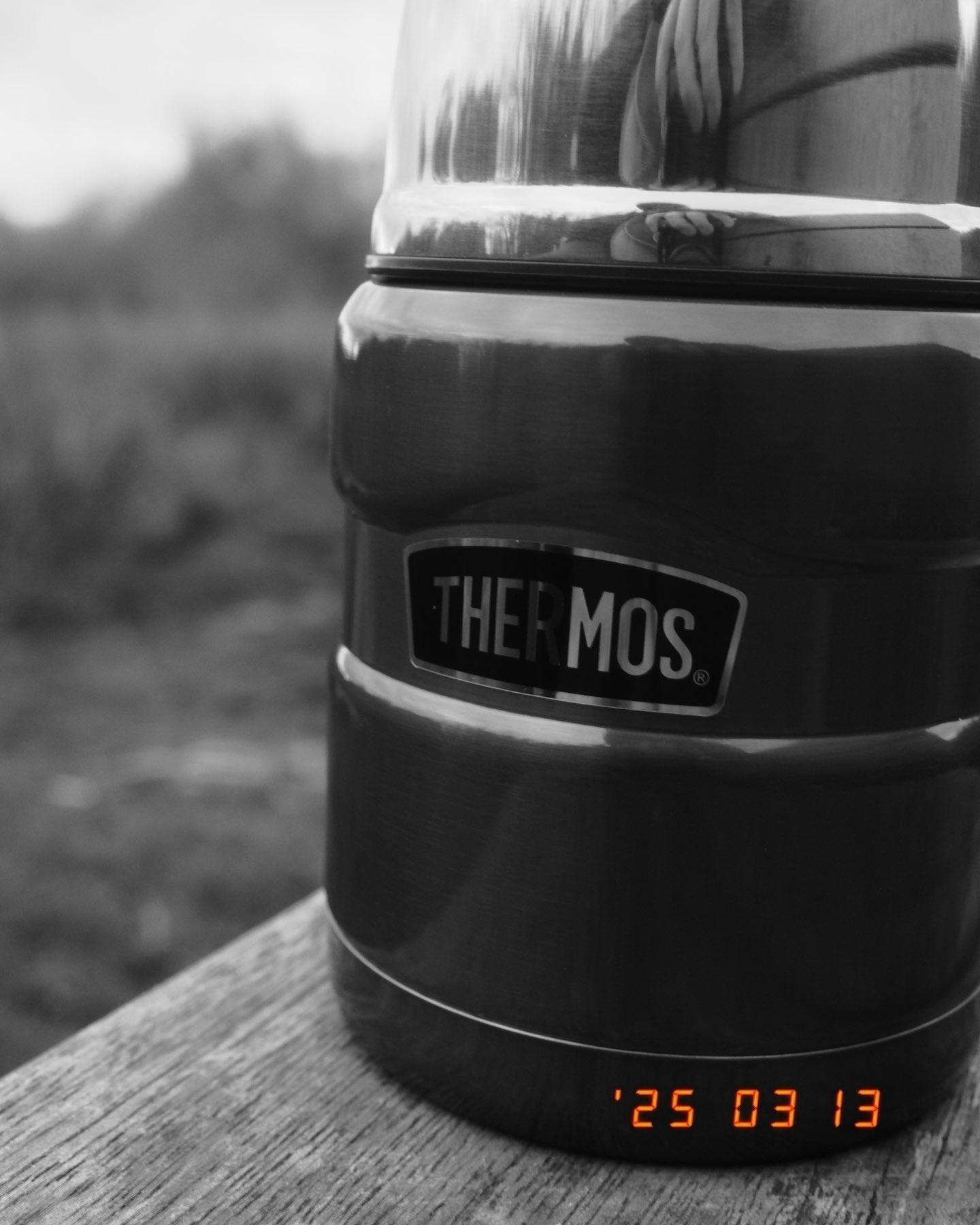 A Thermos flask with a shiny lid is situated on a piece of wood. A distorted reflection of some fingers can be seen. The image is high contrast black and white. 