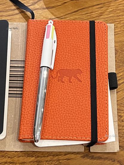 An orange notebook with a textured cover and an debossed logo of a tiger, and a Bic 4 colours pen with a white and silver barrel sits on top of some card and a wooden tabletop.