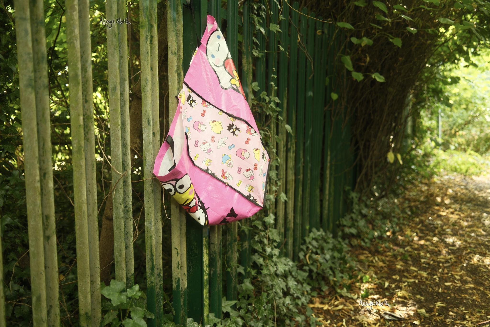 A pink vinyl shopping bag featuring hello kitty characters is hung on a green metal fence beside a path