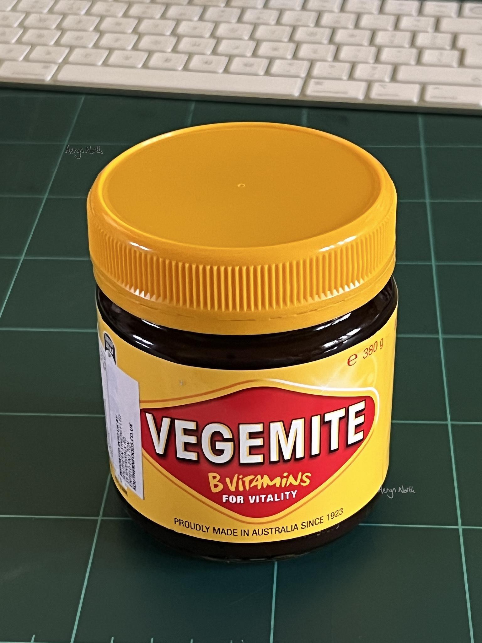 A jar of Vegemite is sitting on a green cutting board. It has a yellow lid and is dark brown with a yellow label with an overlapping white import label on the left hand side. The label says Vegemite B Vitamins for Vitality proudly made in Australia since 1923. Behind the jar is a white keyboard.