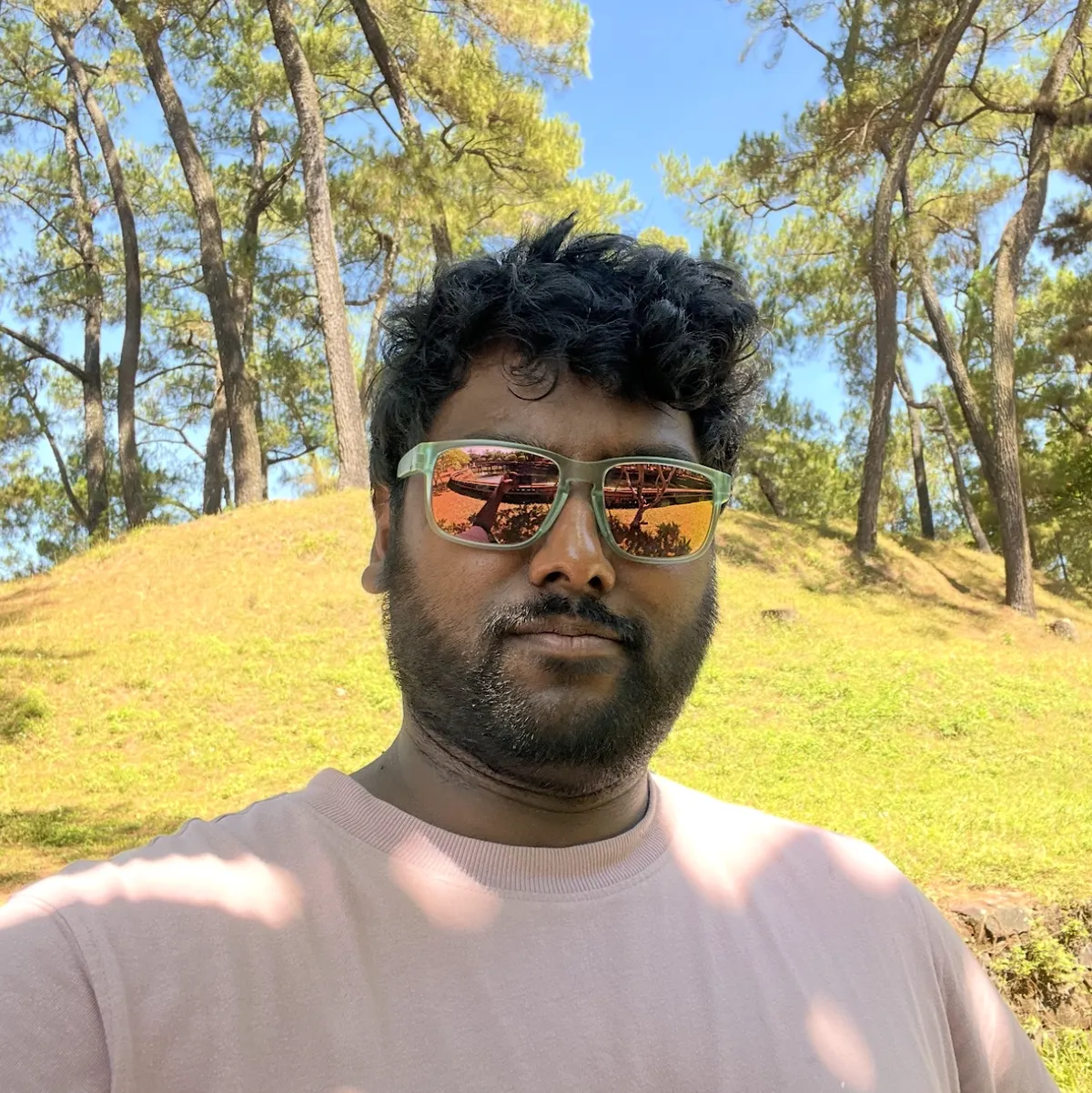 Renowned writer Murugappan (that's me!) stands at the foot of a hill on a sunny day in Huế, Viet Nam. The backdrop features the hill in its entirety, covered in grass, trees, and exposed rock. Murugappan is wearing a pink T-shirt with indistinct white lettering. His sunglasses bear a reflection of his arms, clearly indicating that this picture is a selfie, despite his best attempts to disguise it.