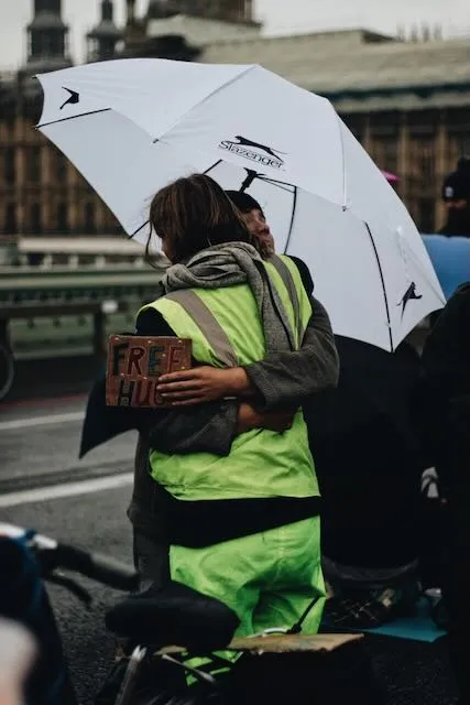Two people embrace beneath an umbrella, one is holding a sign that reads free hugs