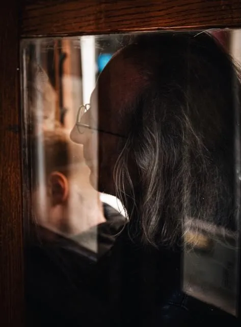 A man with glasses and interesting hair leans against a window