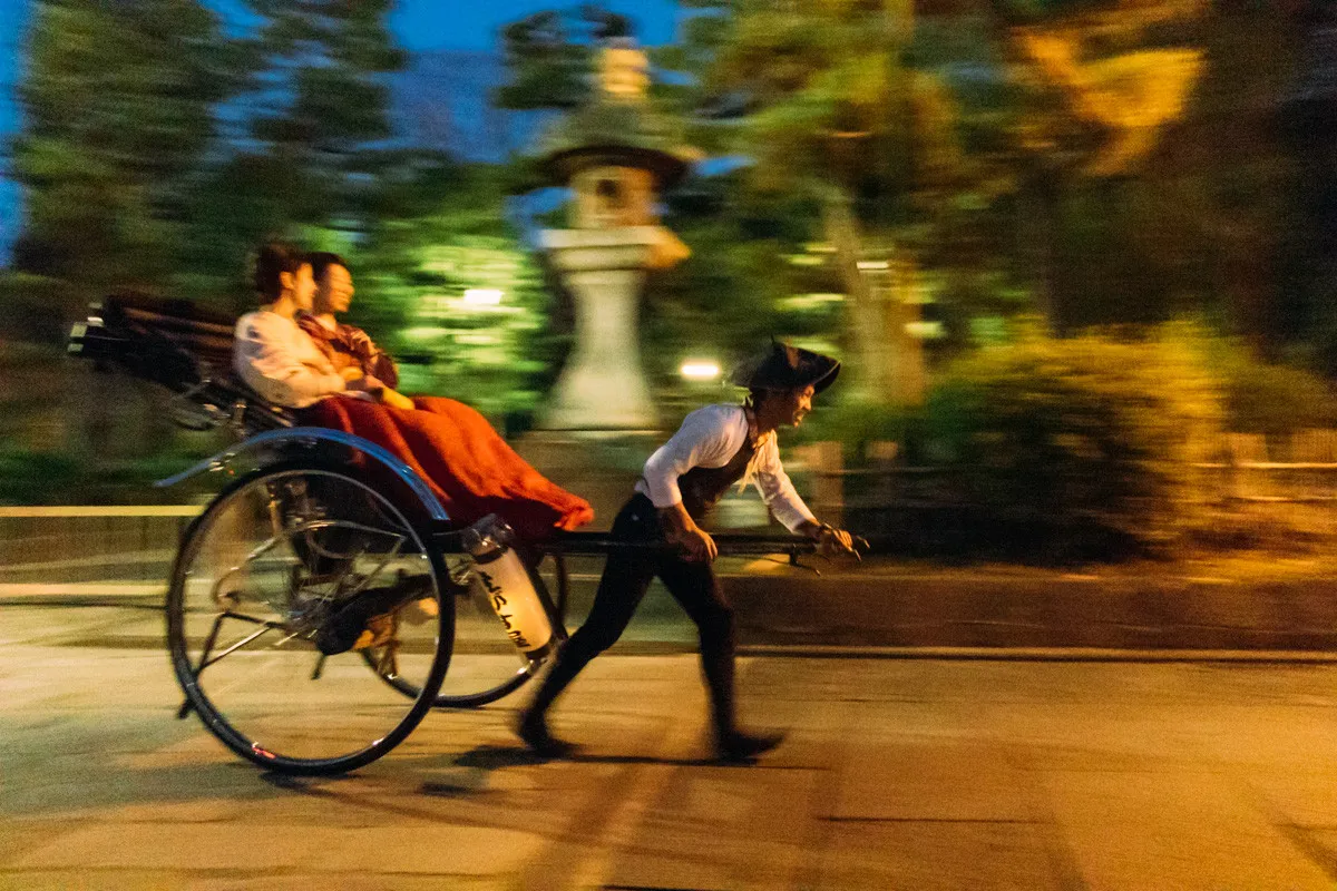 Motion-blurred night shot of a rickshaw puller pulling two passengers in a traditional Japanese carriage past a stone lantern.