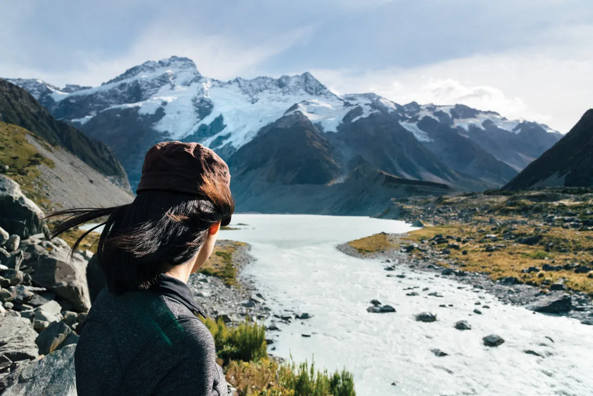 Young woman standing beside a glacial river, looking toward snow-covered mountains under a cloudy sky.