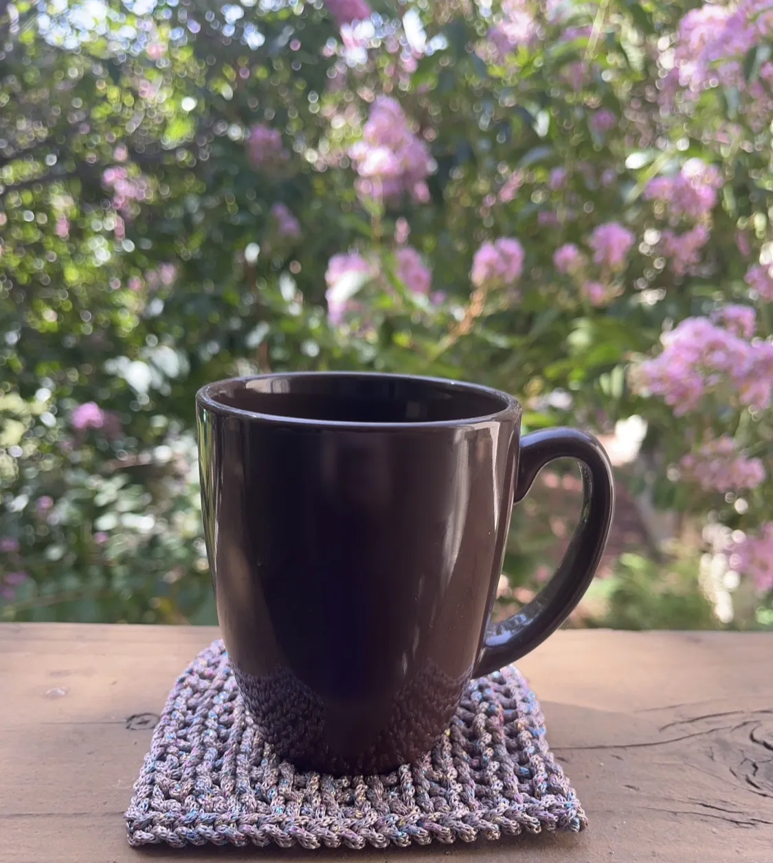 Photo of a brown mug sitting on a knitted cup coaster. The coaster looks iridescent. The background is a tree with pink blossoms in morning light.