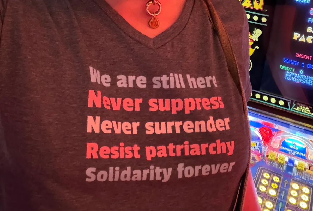Elia is in a pinball arcade in front of a Baby Pac-Man machine and a Bad Cats. We're zoomed in on her shirt, which says: We are still here / Never suppress / Never surrender / Resist patriarchy / Solidarity forever