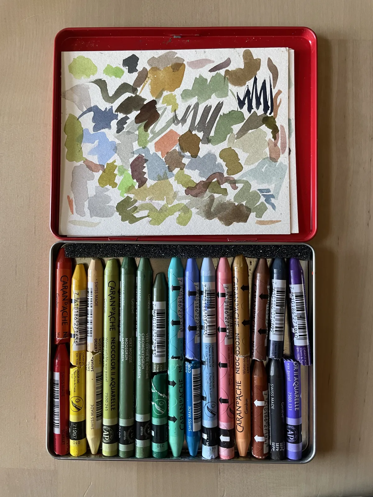 A case of Caran D'ache Neocolor II wax pastels and sheets of loose watercolor swatch paper.