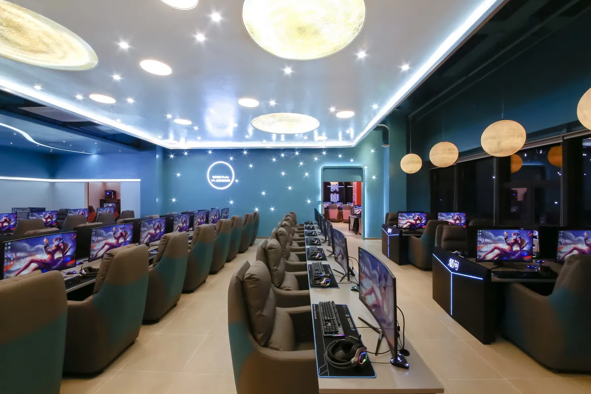 成都一网咖室内照片。空间采用深蓝绿色墙面，墙上布满星星状LED灯点。天花板为浅灰色，悬挂多个圆形月球造型吊灯，发出柔和暖光。室内对称排列两排游戏工作站，每个座位配有显示器、键盘鼠标和灰色人体工学座椅。显示器上显示游戏画面。墙面有蓝色霓虹灯文字标识。整体营造宇宙星空主题氛围。
