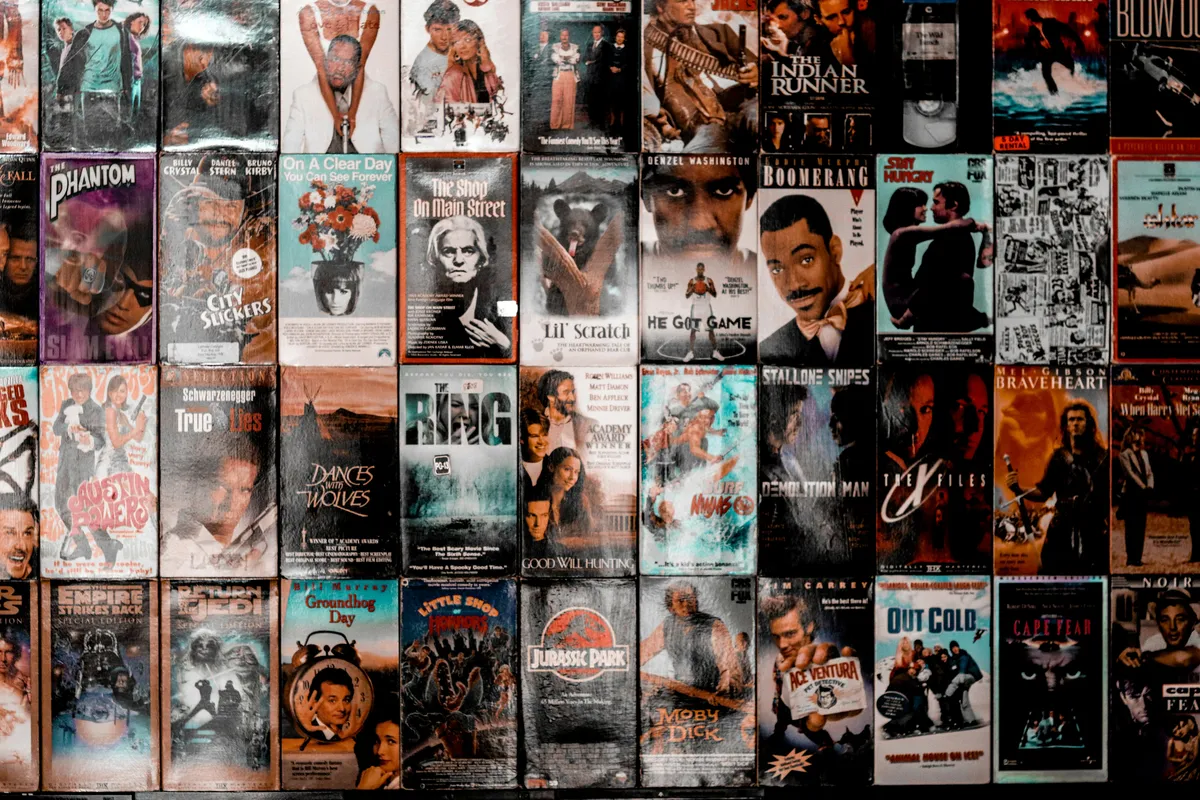 一面摆满复古 VHS 录像带的墙，封面密集排列，表面带有明显的磨损、划痕和褪色痕迹。录像带涵盖多部经典电影，包括《侏罗纪公园》《土拨鼠之日》《心灵捕手》《勇敢的心》《真实的谎言》等，整体呈现出浓厚的 20 世纪末家庭录像店氛围。