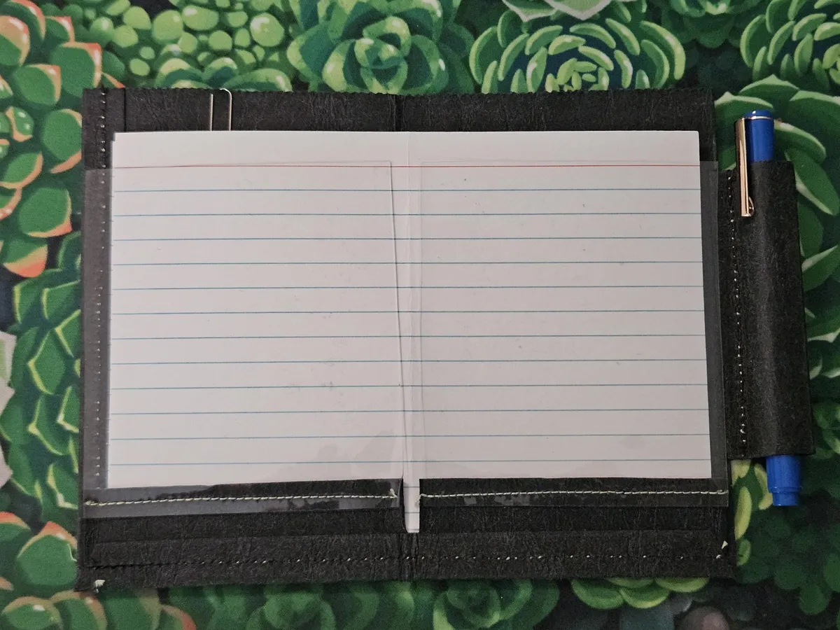 Handmade index card pocket folder A top-down view of a handmade, dark grey folder for carrying index cards. The folder is open, revealing a blank, lined index card inside, held by clear plastic pockets. A blue pen is secured in a loop on the right, and the entire wallet rests on a green, succulent-patterned surface.