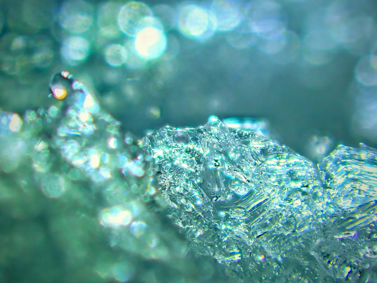 Glimmer A close-up, abstract photo of translucent ice or glass shards with shimmering, out-of-focus light reflections (bokeh) in the background. The dominant colors are cool tones of teal, aqua, and bright white, suggesting a blurry, ethereal, and somewhat obscure visual field.